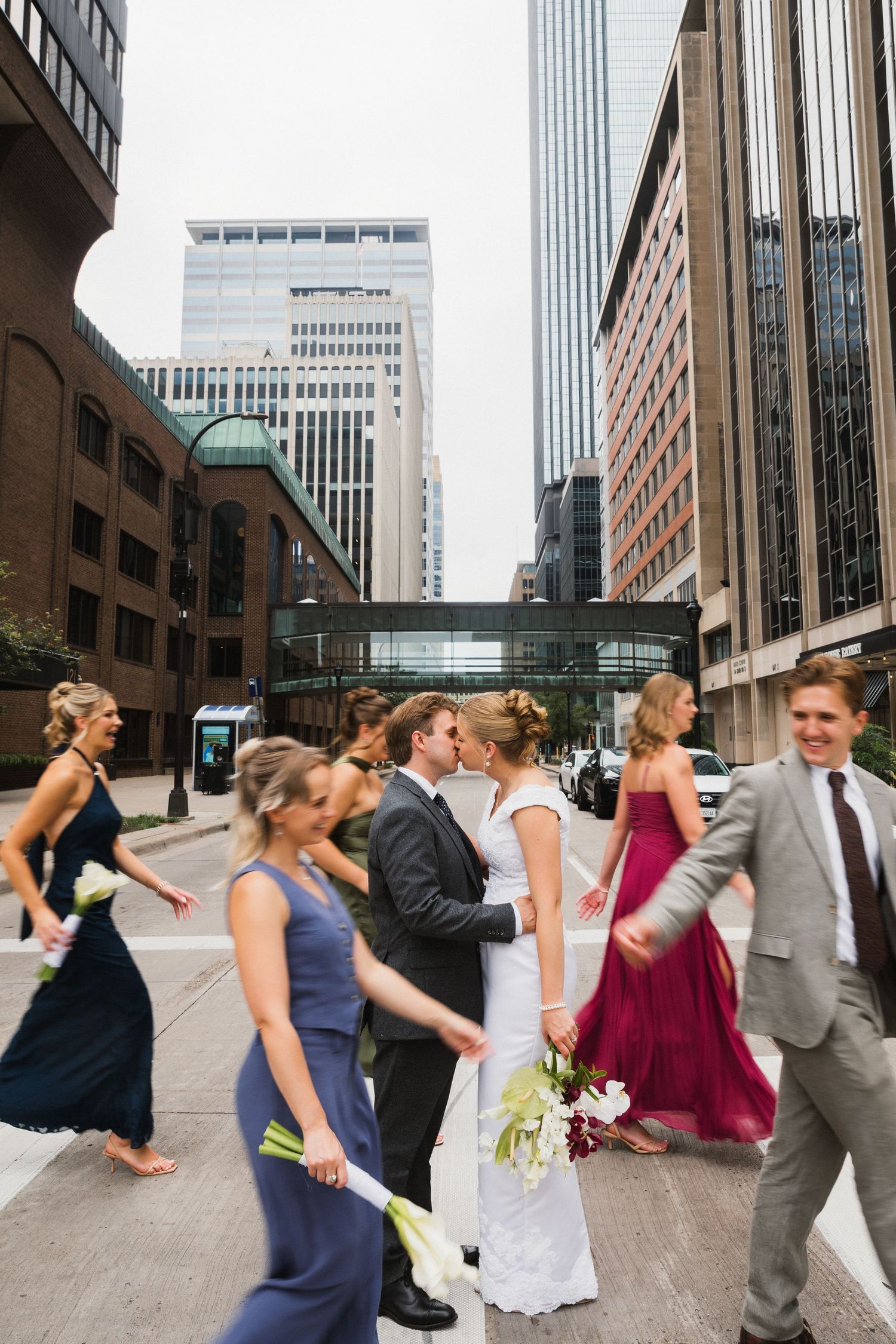 A bride and groom share a kiss while crossing a city street surrounded by bridesmaids in navy and burgundy gowns.