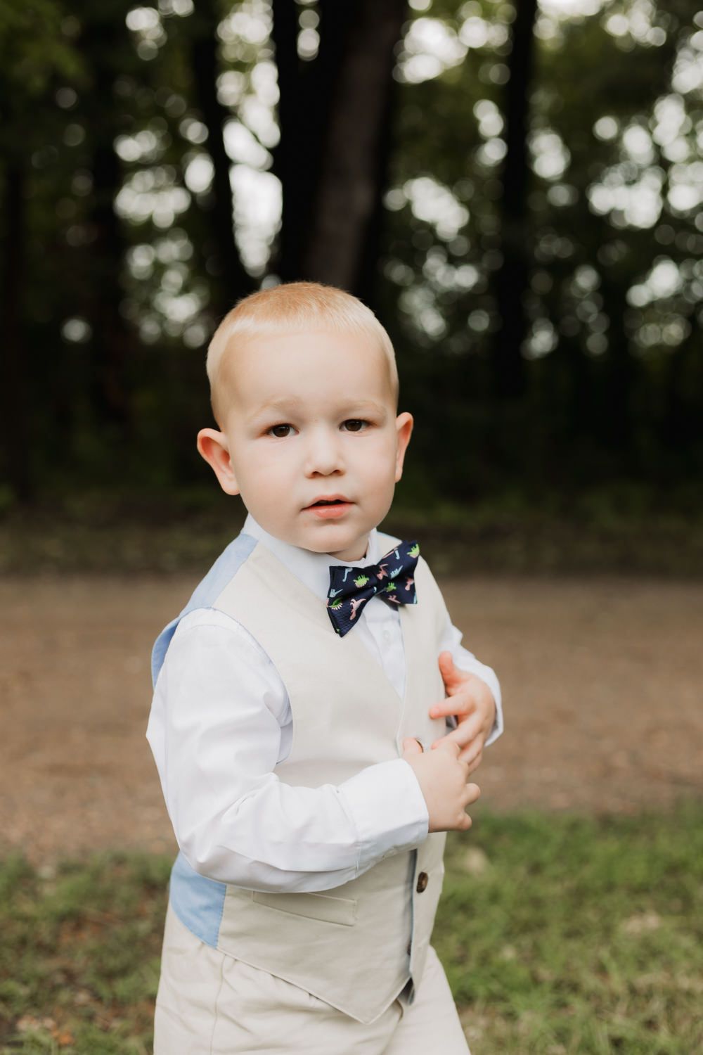 A small child in formal attire with a bowtie stands outdoors in a white dress shirt and light colored vest.