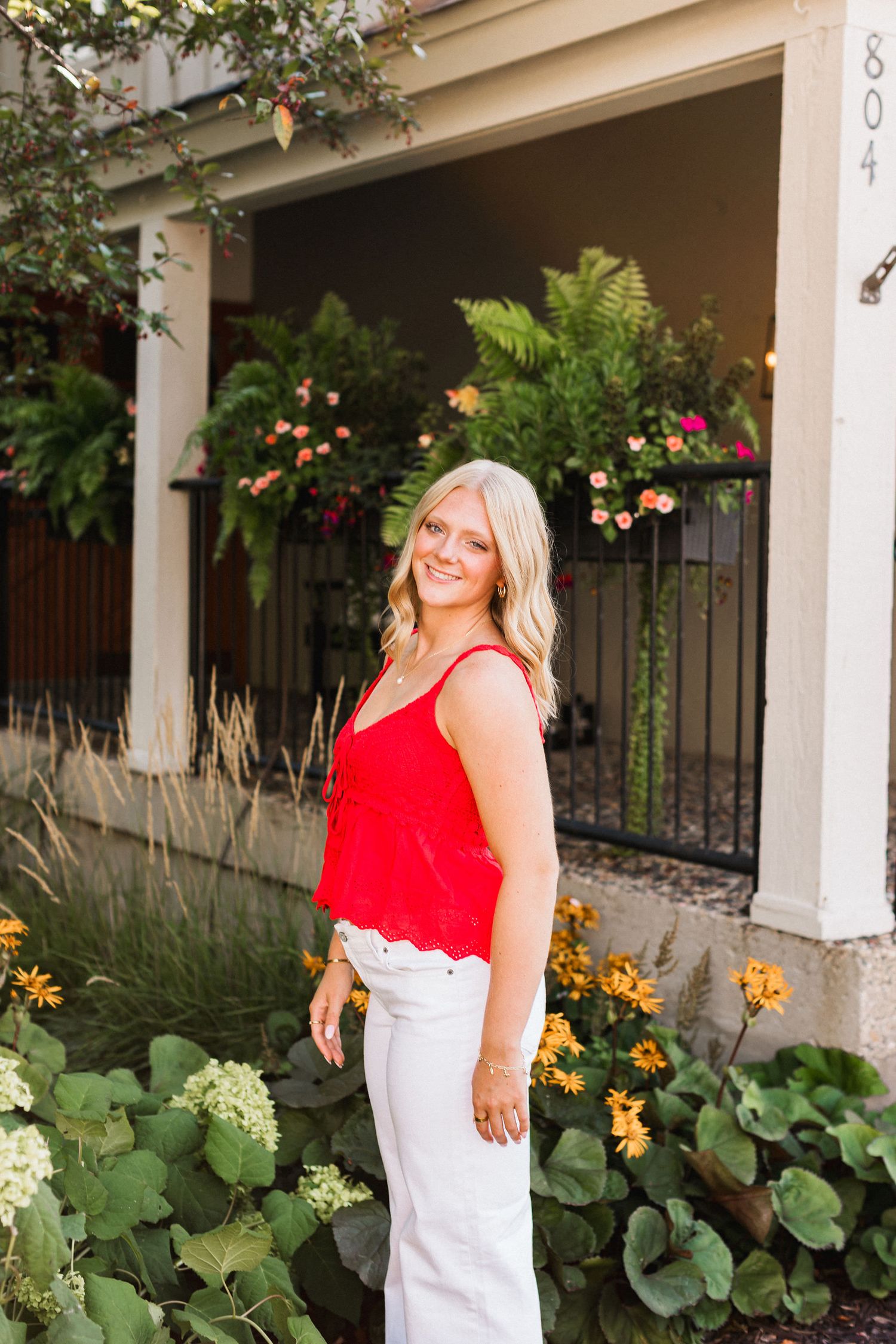 A bright red sleeveless top paired with white pants stands out against a garden backdrop with blooming flowers.