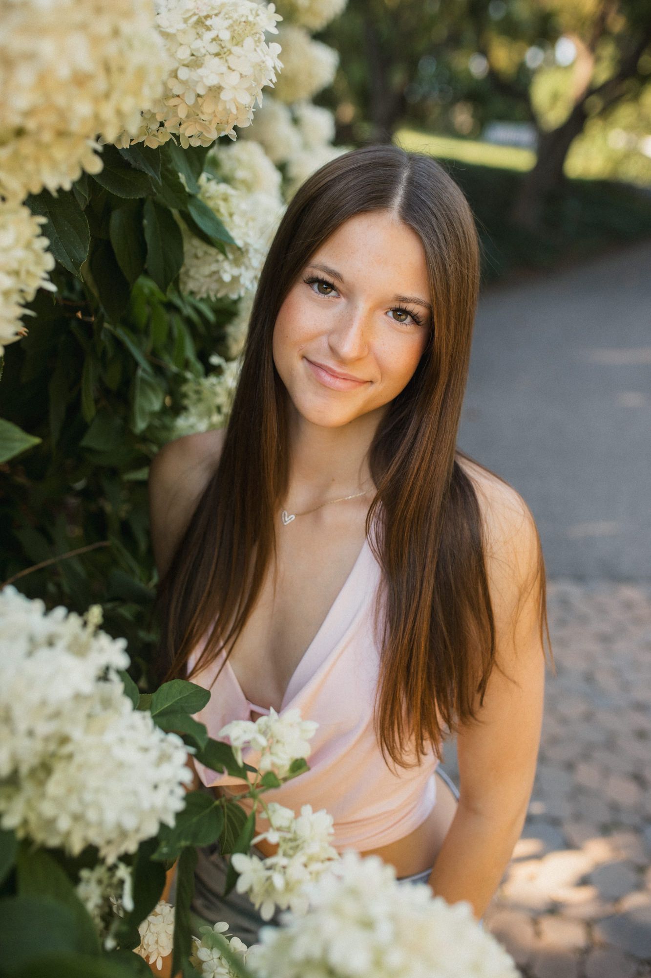 Young woman with long brown hair smiling among white hydrangea bushes in a garden wearing a pink dress.