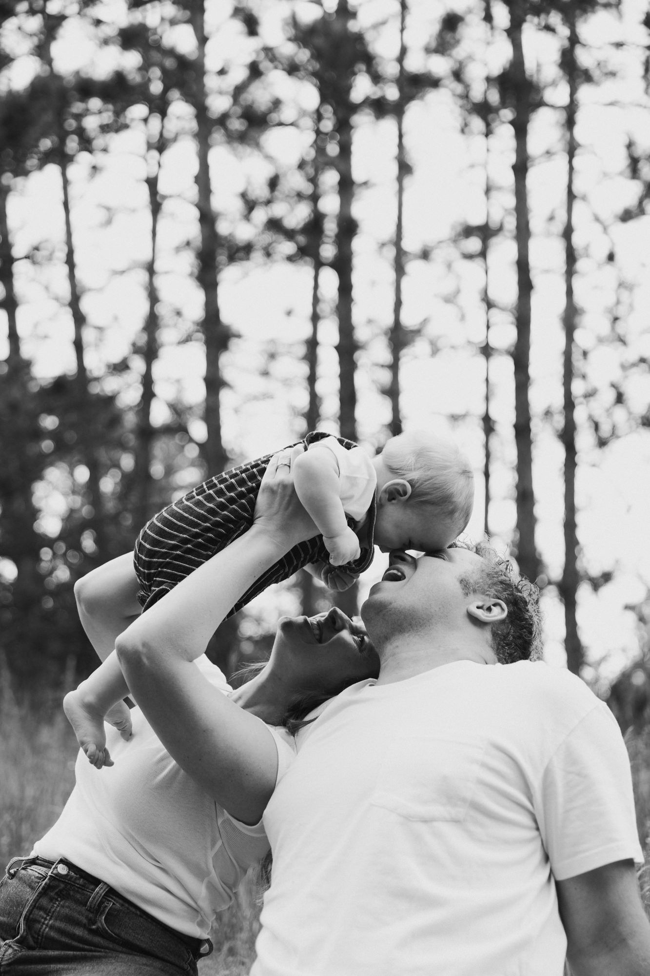 Black and white photo of two parents lovingly lifting their baby up for a kiss outdoors among tall trees.