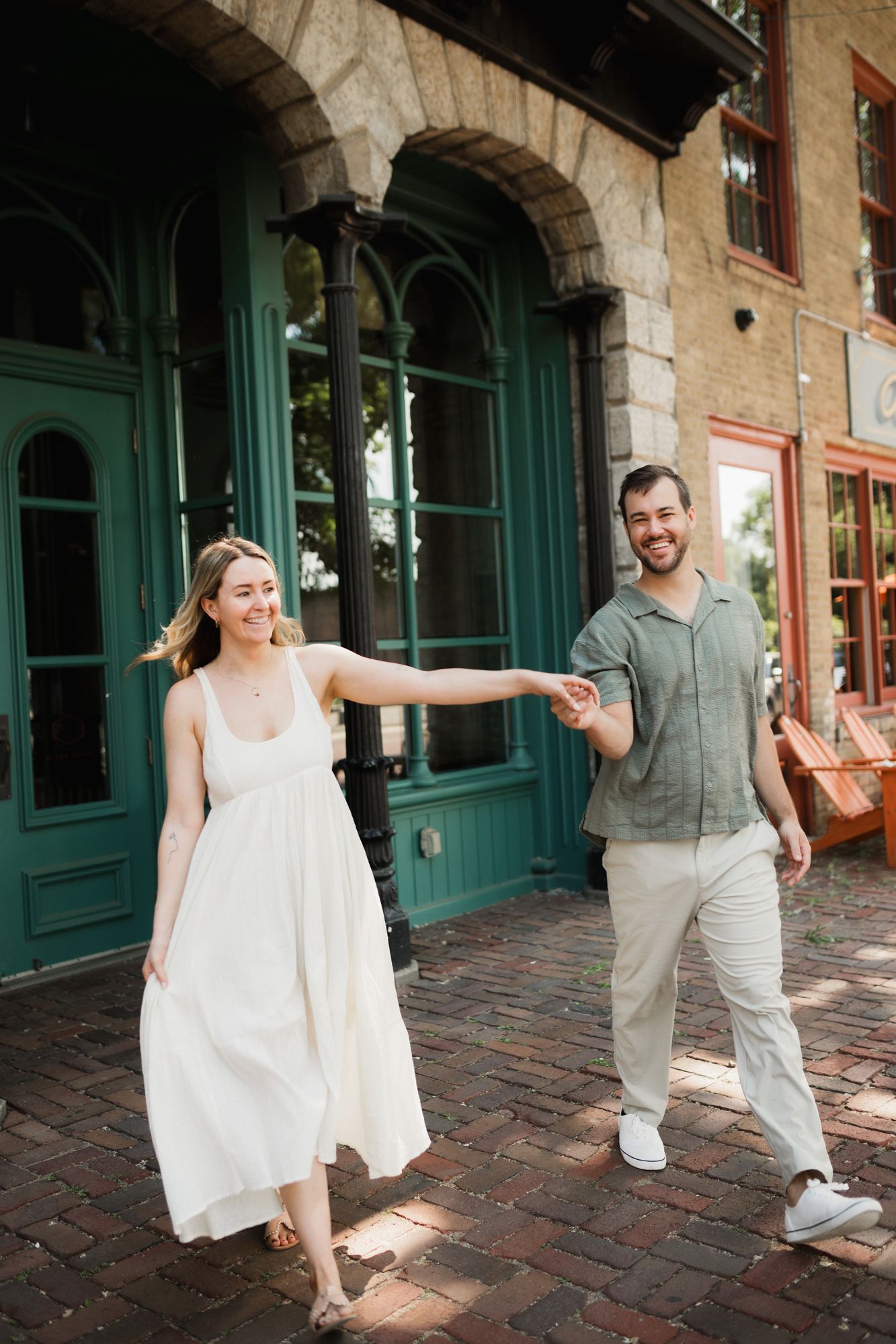 A smiling couple holds hands walking on a brick path outside a building with green doors.