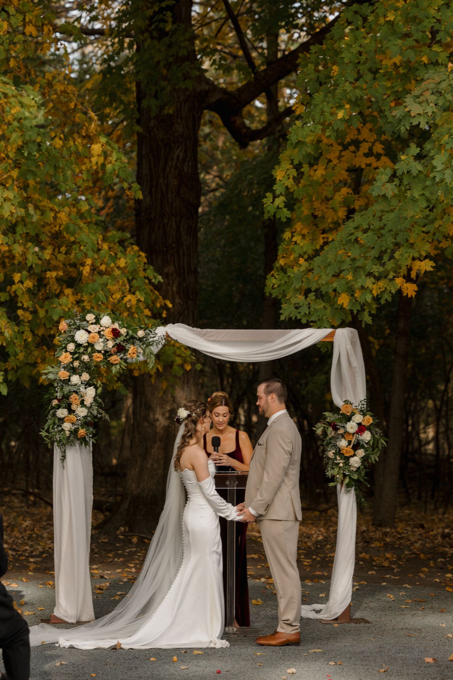 Bride and groom hold hands during an outdoor autumn wedding ceremony beneath a floral draped arch with fall foliage behind.