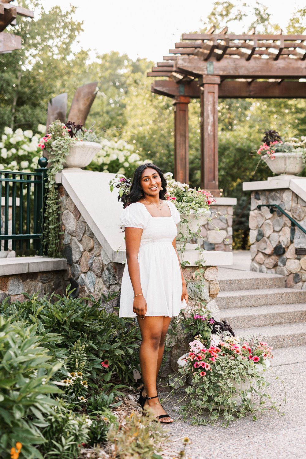 White dress against stone wall and wooden pergola in a garden with blooming flowers.