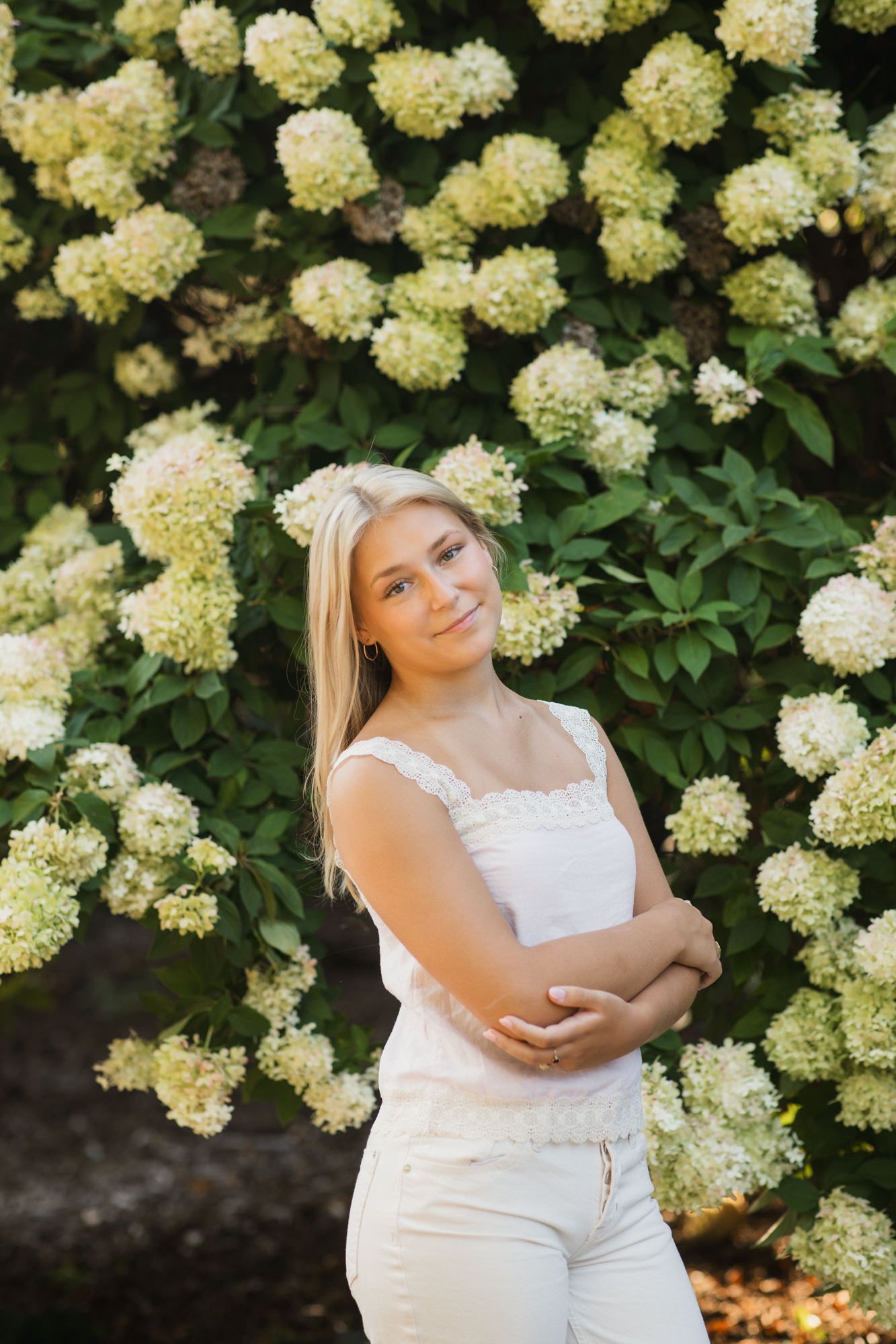 Young blonde woman in white top and jeans poses with arms crossed before a lush hydrangea bush in bloom.