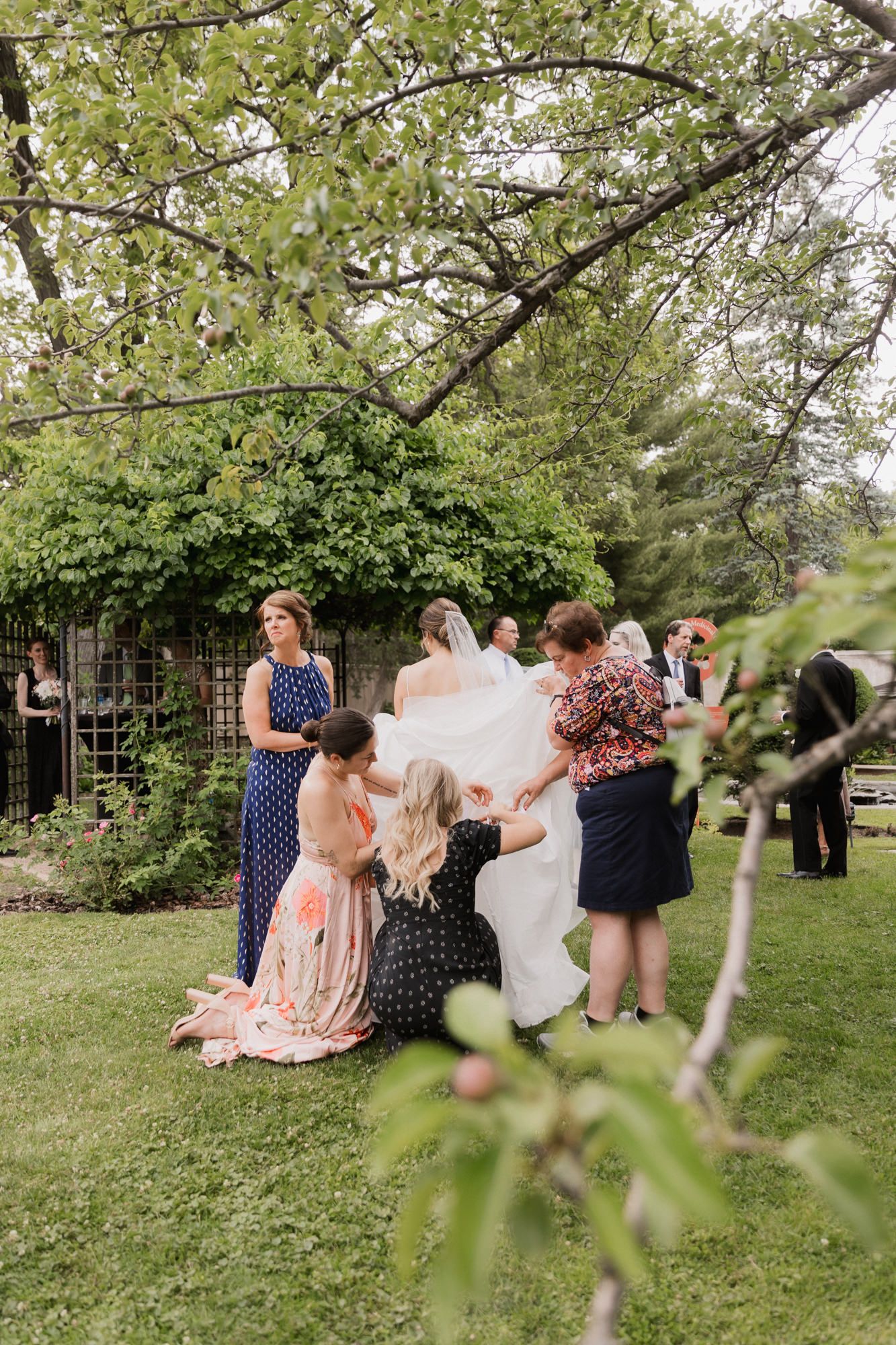 Bridesmaids helping bride with wedding dress train in lush garden setting during outdoor wedding ceremony.