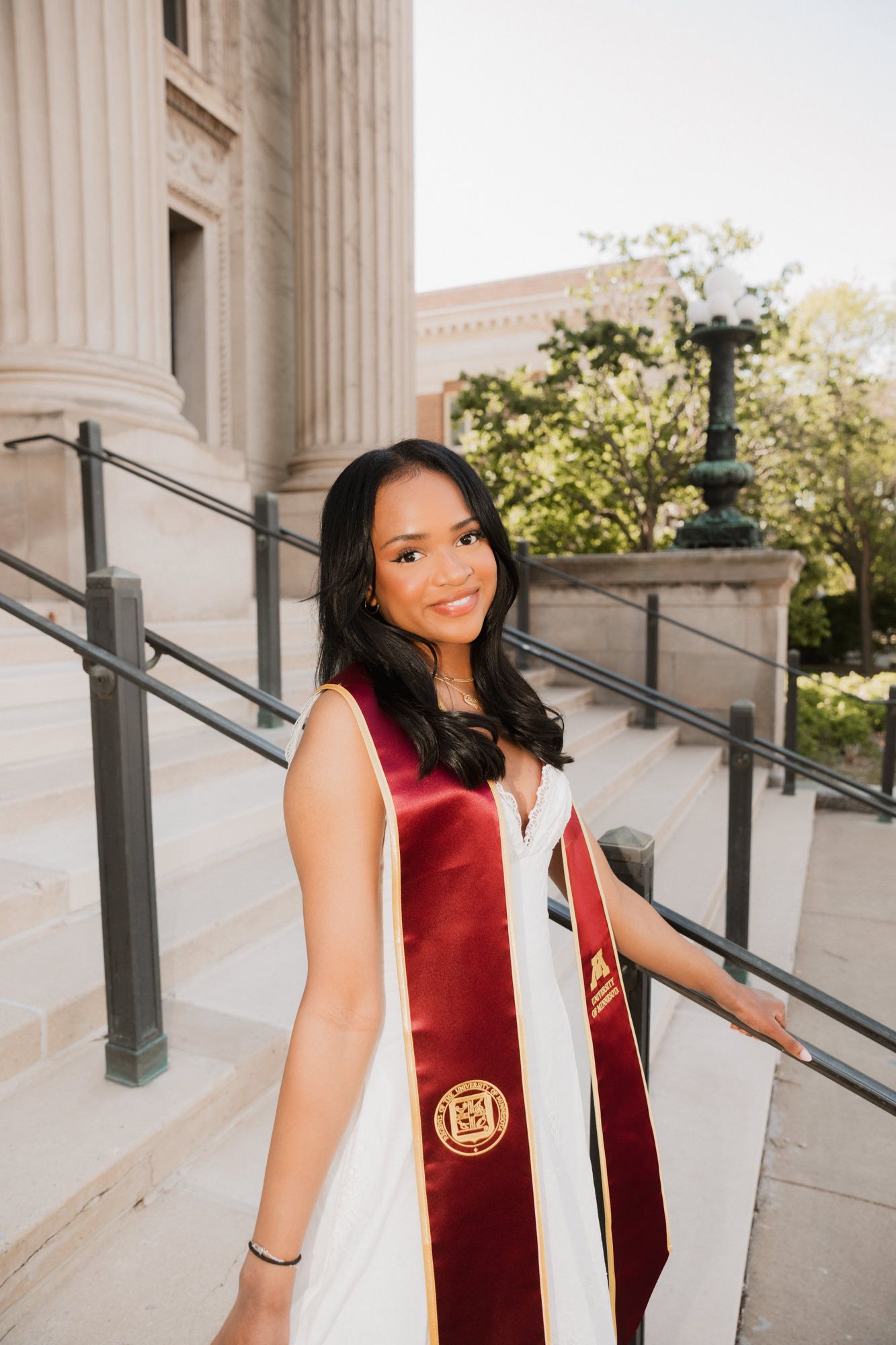 Young woman in white dress and maroon graduation stole smiles on university steps during commencement photo session.