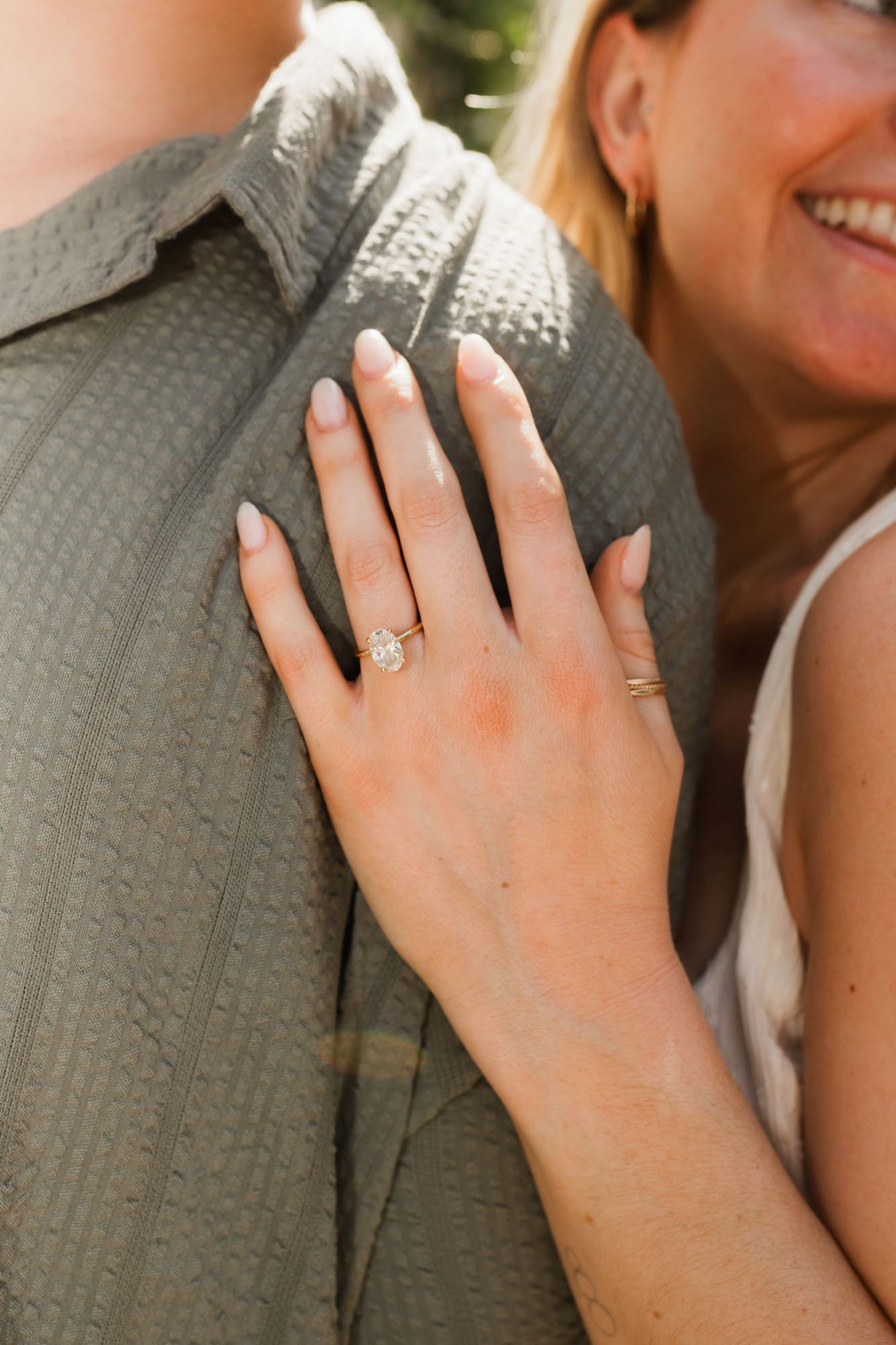 Close-up of a woman's hand displaying a stunning oval diamond engagement ring while resting on a man's shoulder.