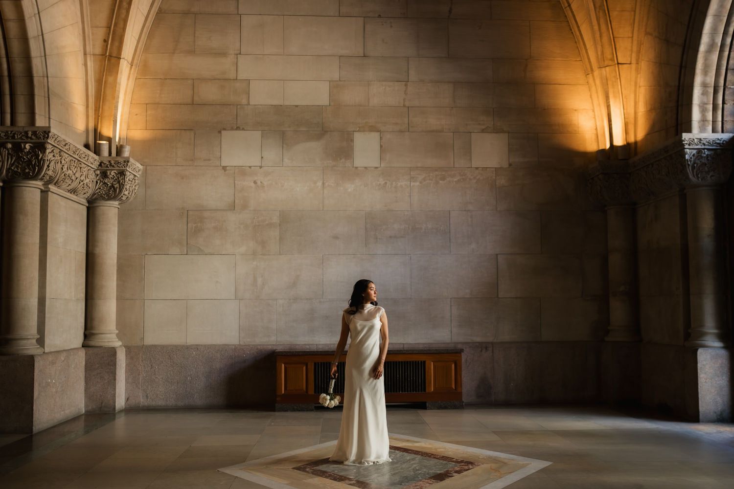 Bride in white gown stands in grand stone cathedral hall with arched columns and warm ambient lighting.