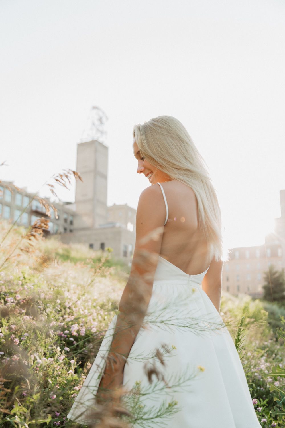 A person in a flowing white dress stands in a sunlit garden with wildflowers and urban buildings in the background.