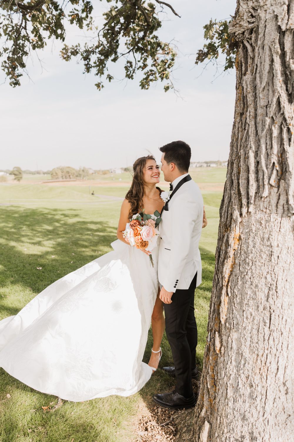 Romantic wedding couple poses between large trees in a scenic outdoor setting with flowing white dress.