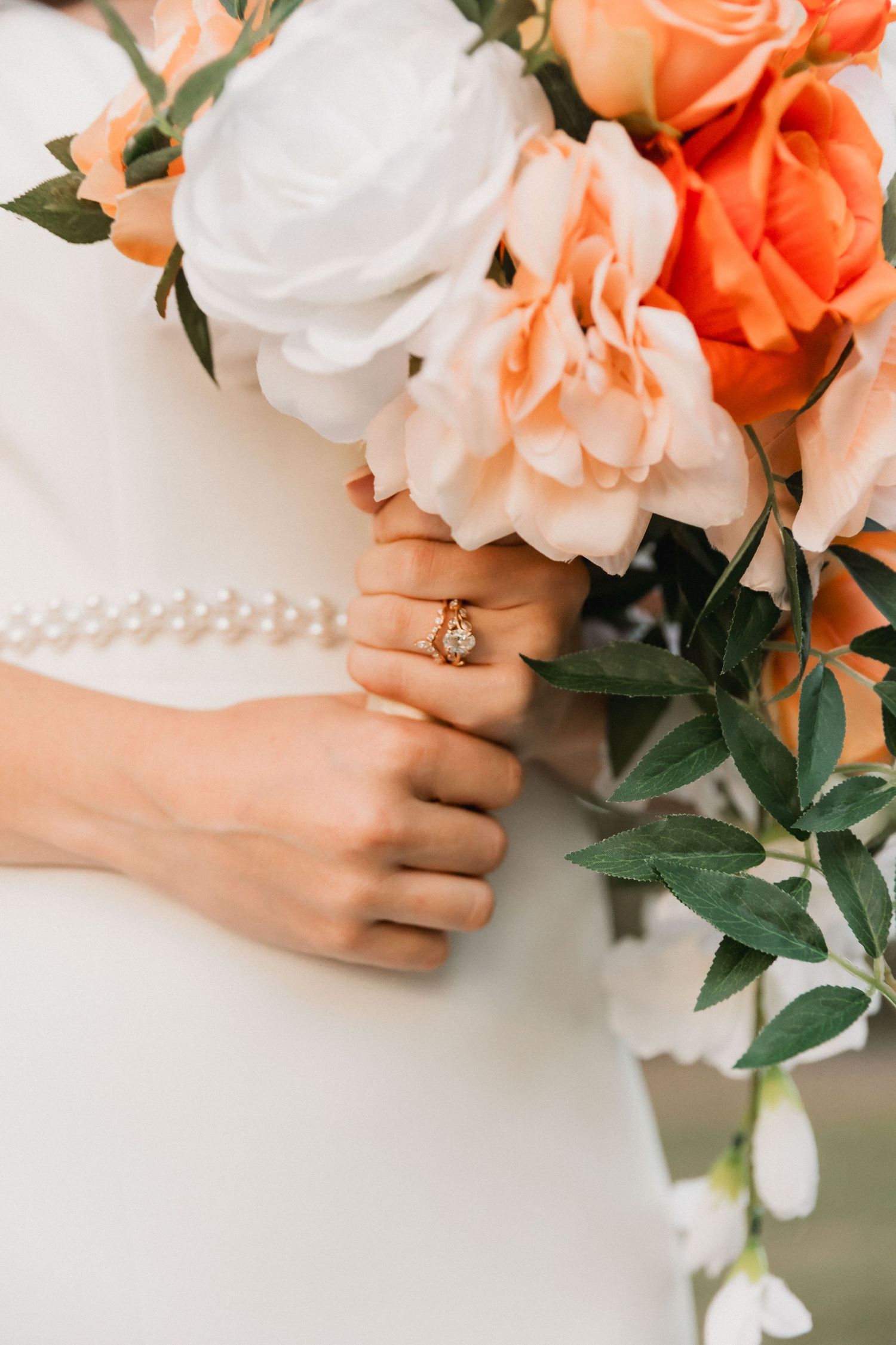 A hand wearing an engagement ring holds a romantic bouquet of coral and white roses with green foliage.