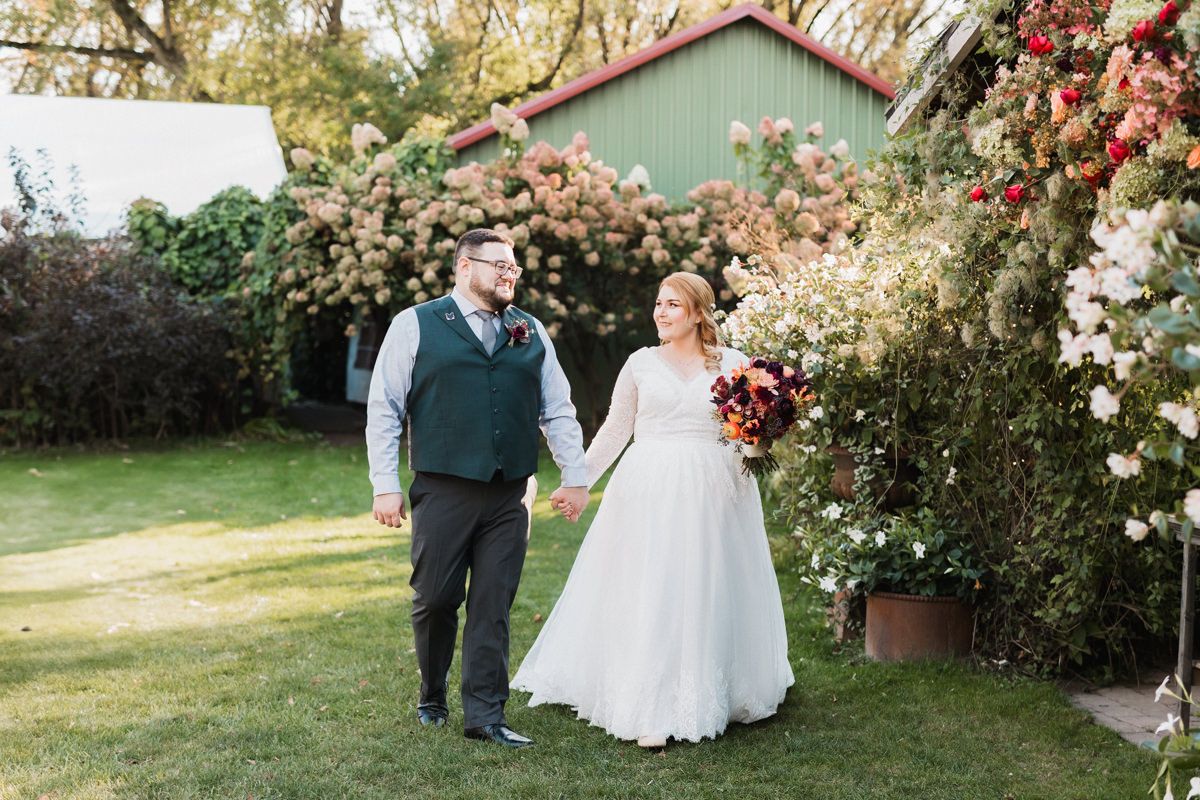 Wedding party members stroll through a garden path with roses and a barn in the background.
