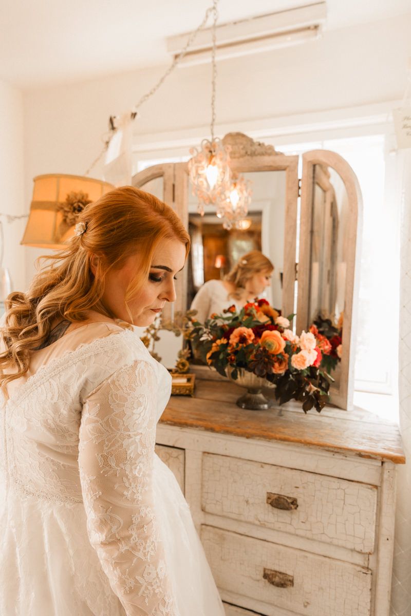 A bride in a long-sleeved lace wedding dress stands in front of a vintage white vanity with floral arrangements.