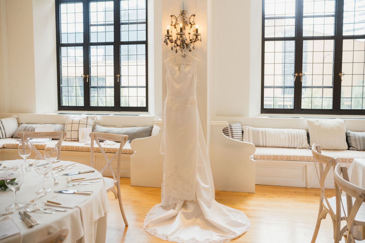 White wedding dress hanging near chandelier in elegant reception room with wooden floors and white-draped tables.