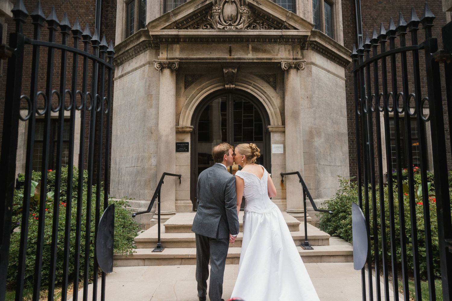 Bride and groom kissing in front of elegant stone building entrance, framed by ornate black iron gates.