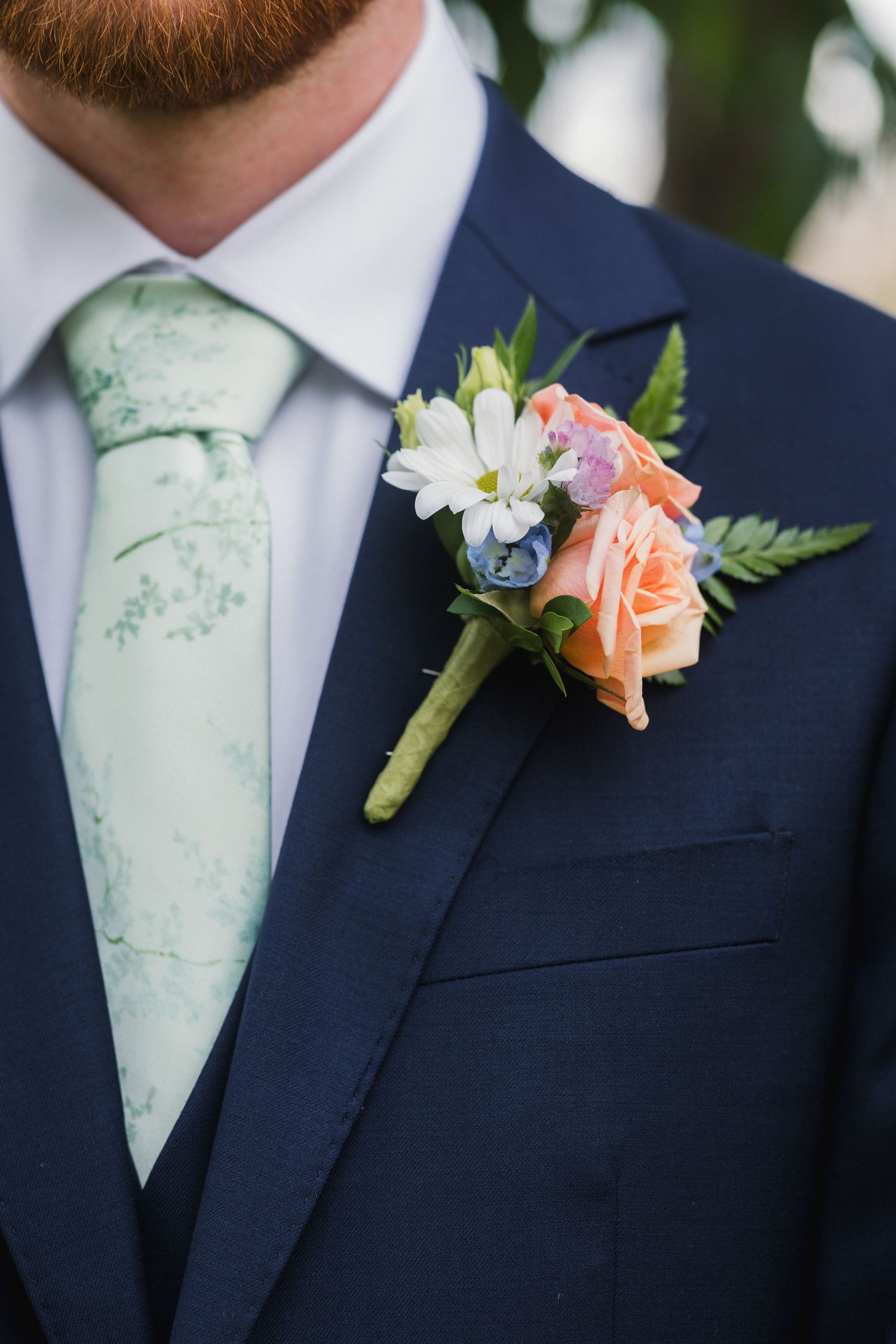 Groom in navy suit with mint floral tie and peach rose boutonniere at wedding.