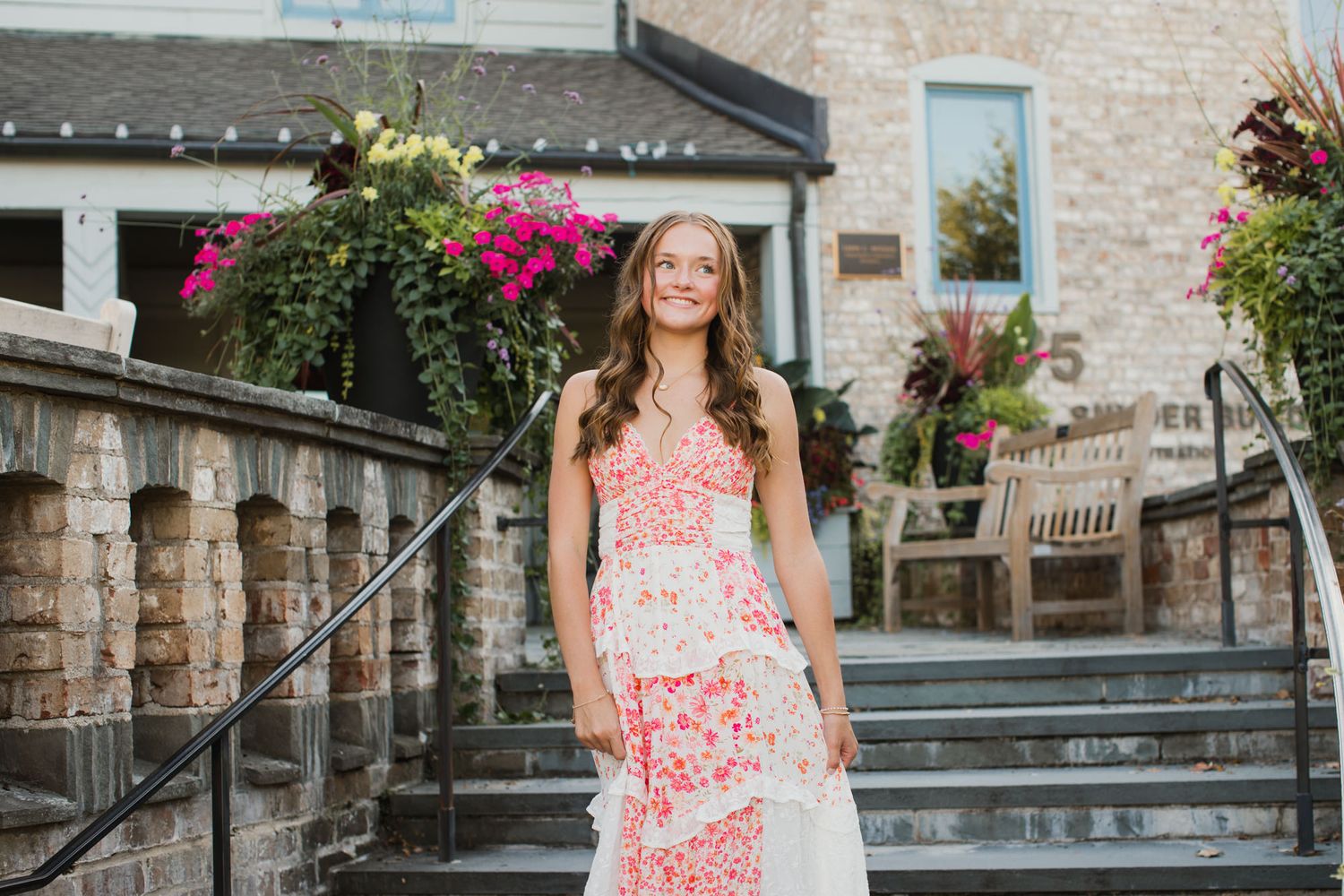 Young woman in a floral tiered maxi dress smiling on stone steps outside a building with pink flowers.
