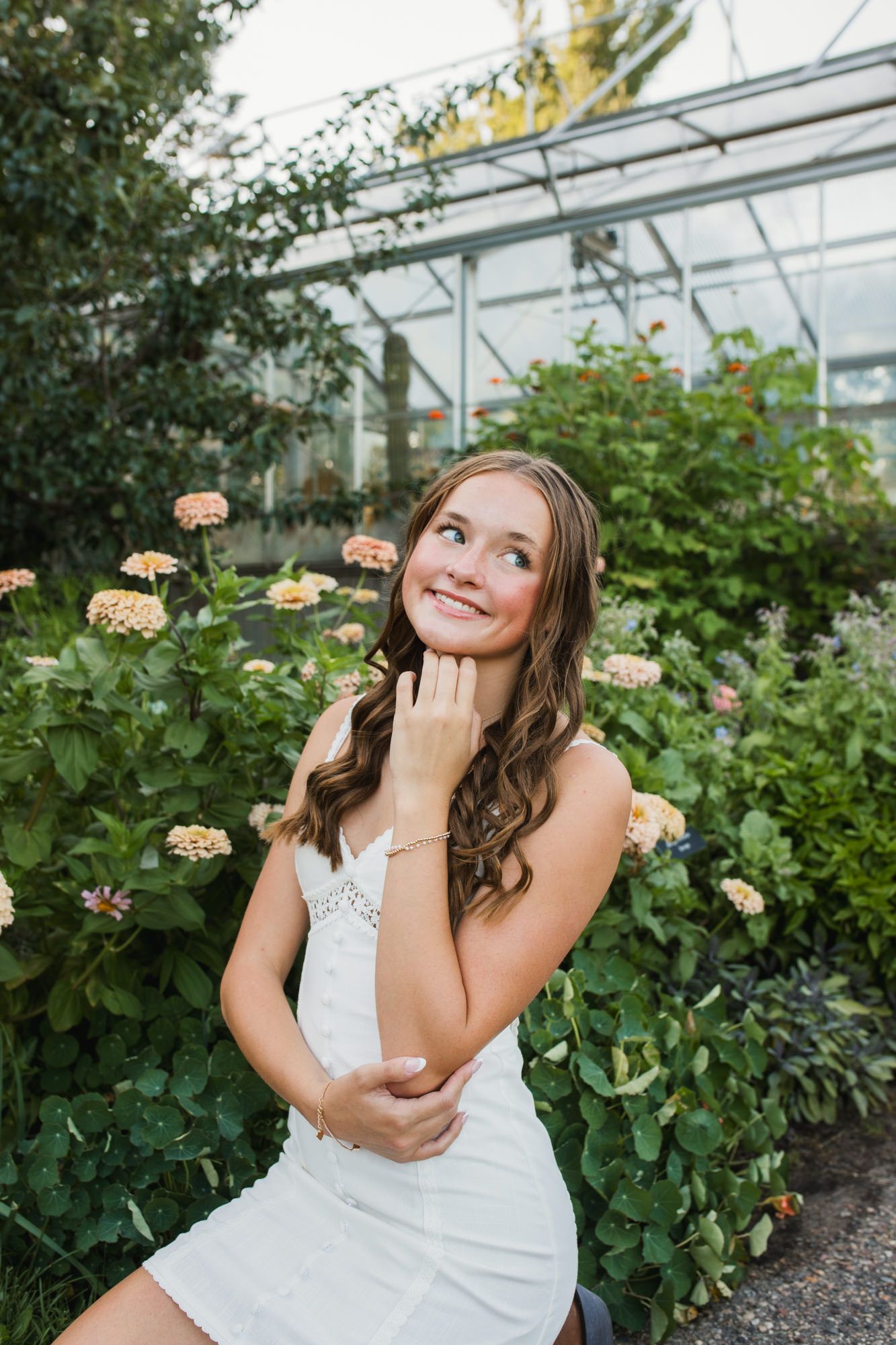 Young woman in white dress smiling in a lush greenhouse surrounded by blooming flowers.
