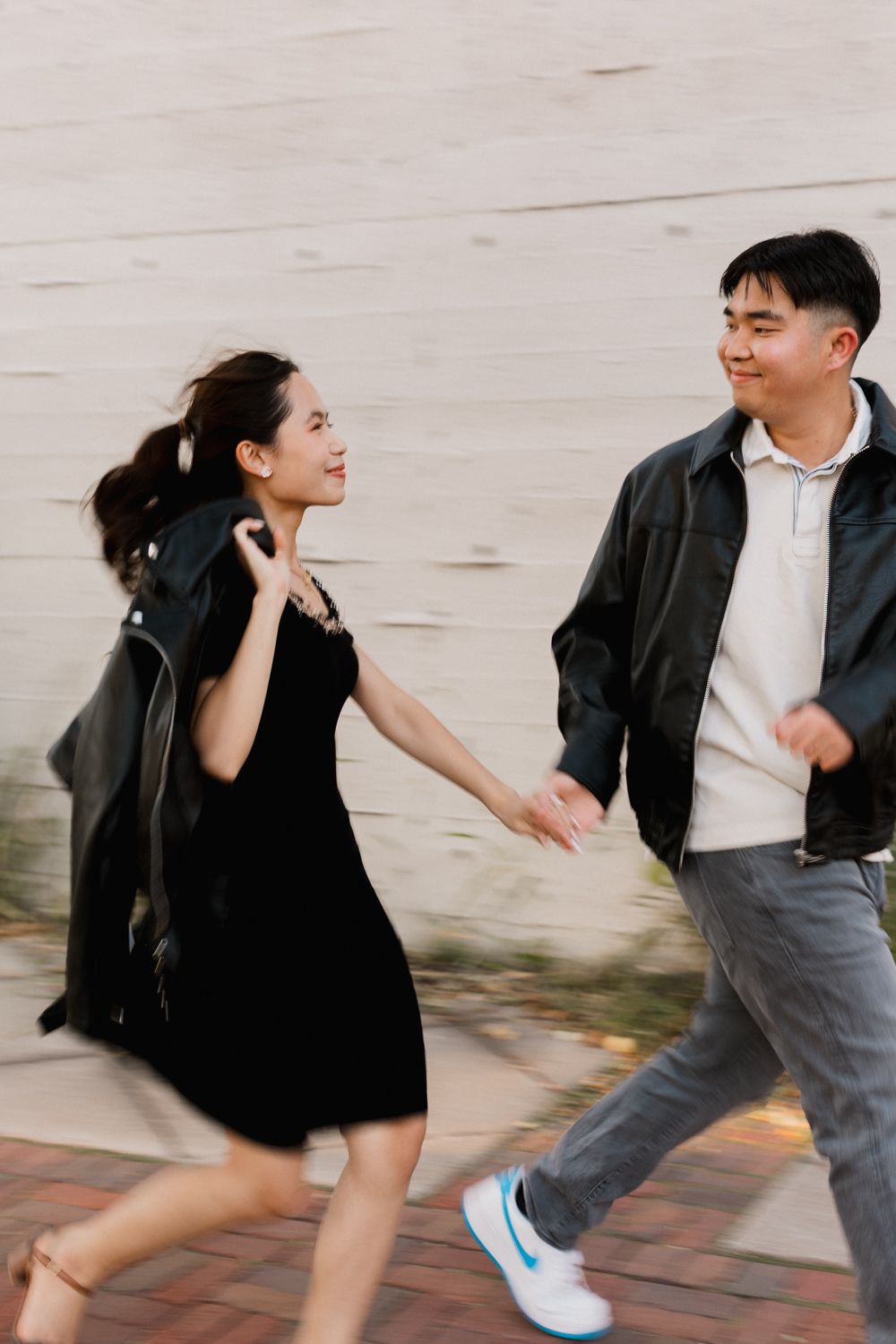 Two people in casual attire run playfully together on a paved walkway against a light-colored wall.
