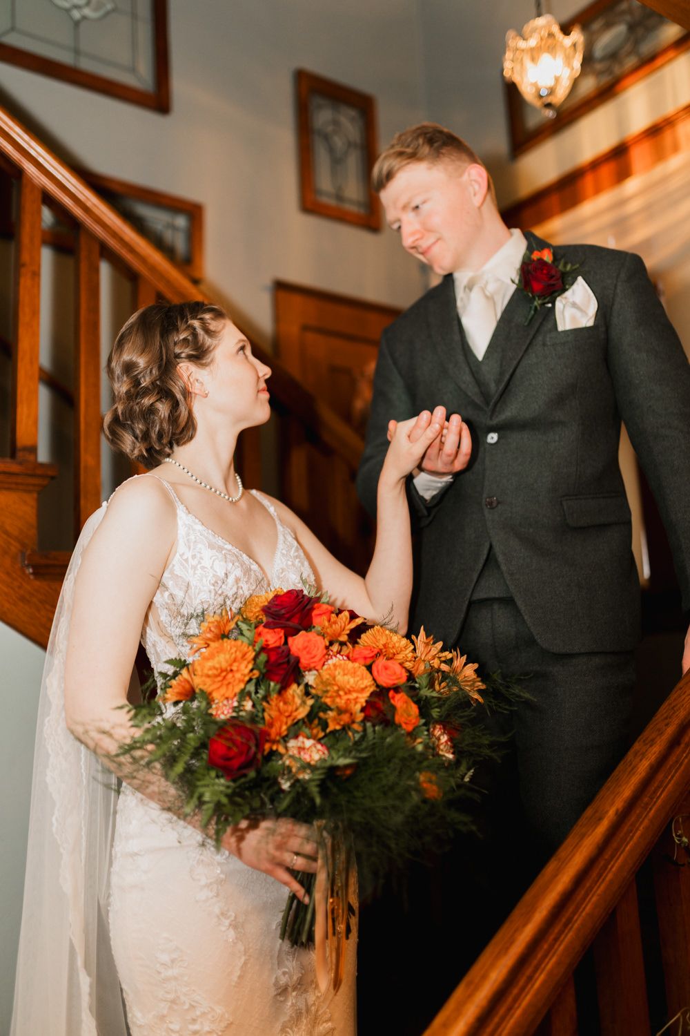 A romantic wedding moment captured on a wooden staircase with orange and red floral bouquets.