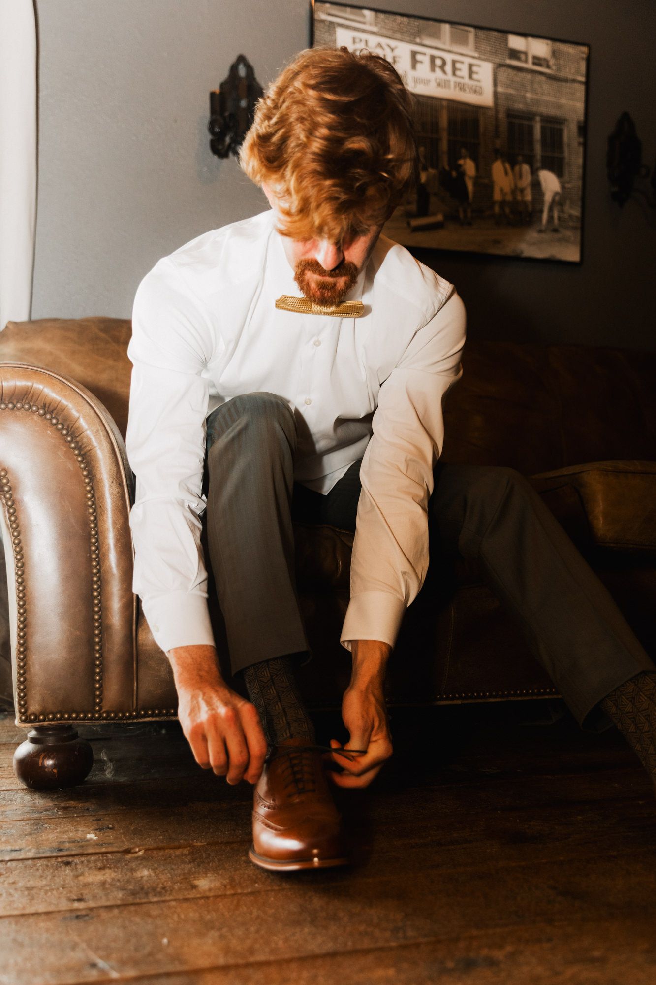 A well-dressed redhead man in white shirt and bow tie tying brown dress shoes while seated on a leather couch.
