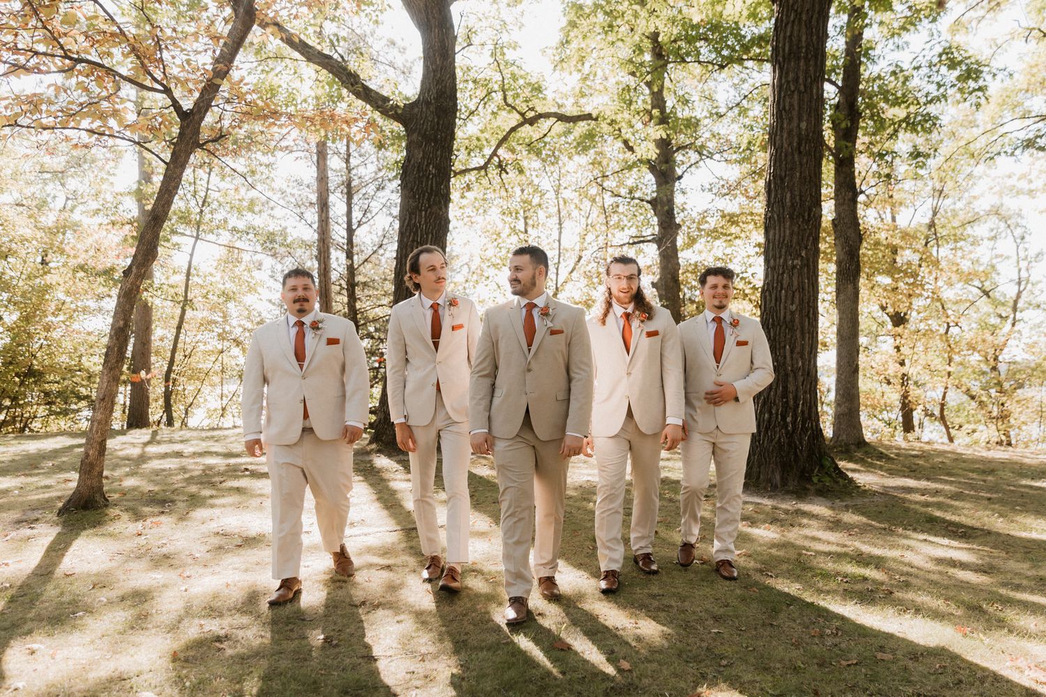 Groom and groomsmen in tan suits with burgundy boutonnieres walking outdoors among trees on a sunny wedding day.