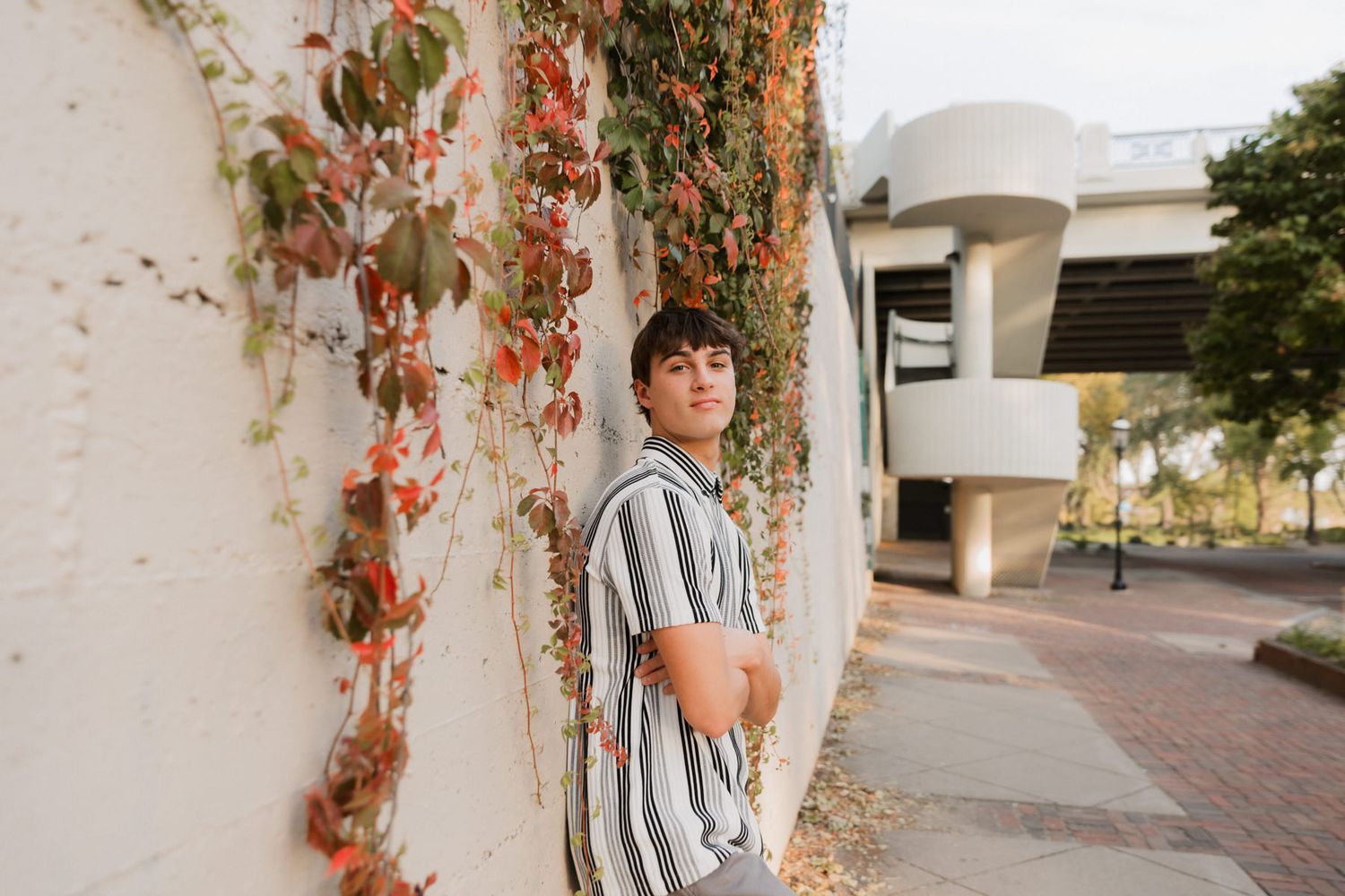 Young man in striped shirt leaning against white wall with red ivy in urban outdoor setting.
