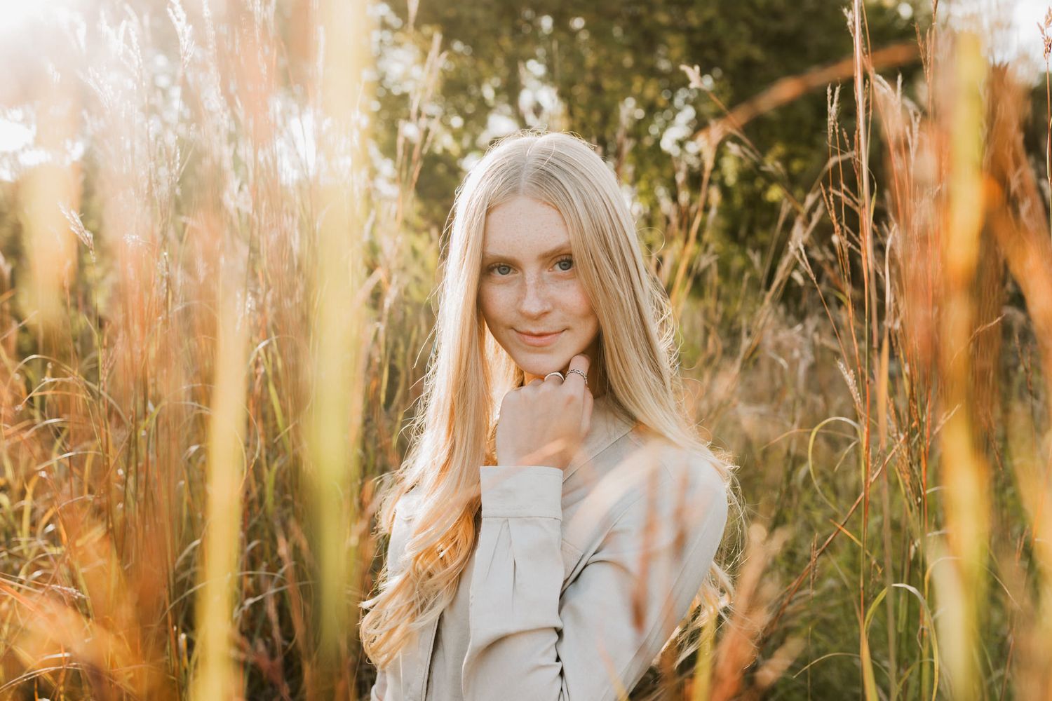 Young blonde woman in beige jacket posing among tall golden grass in soft natural sunlight portrait.