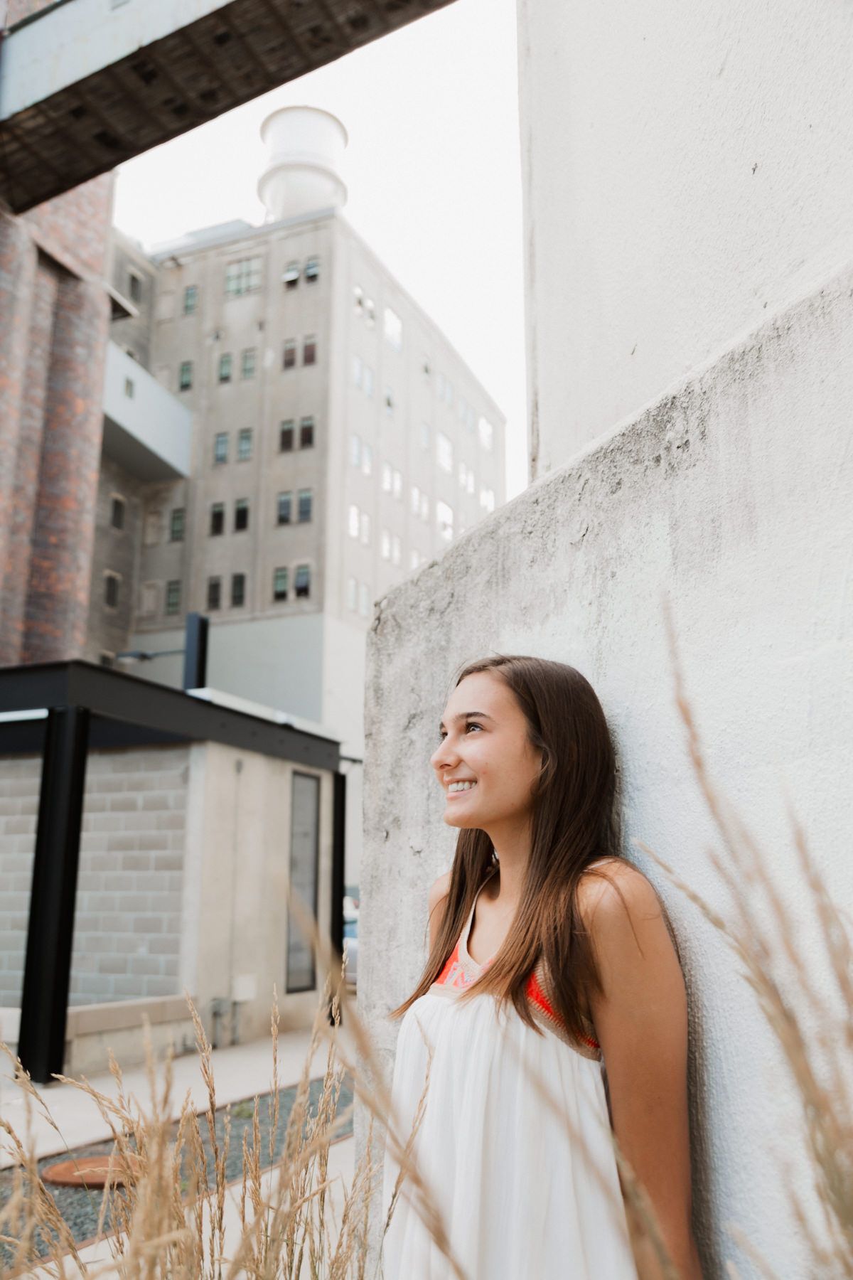 Young woman in white dress smiling while leaning against a white wall in an urban setting with tall buildings.
