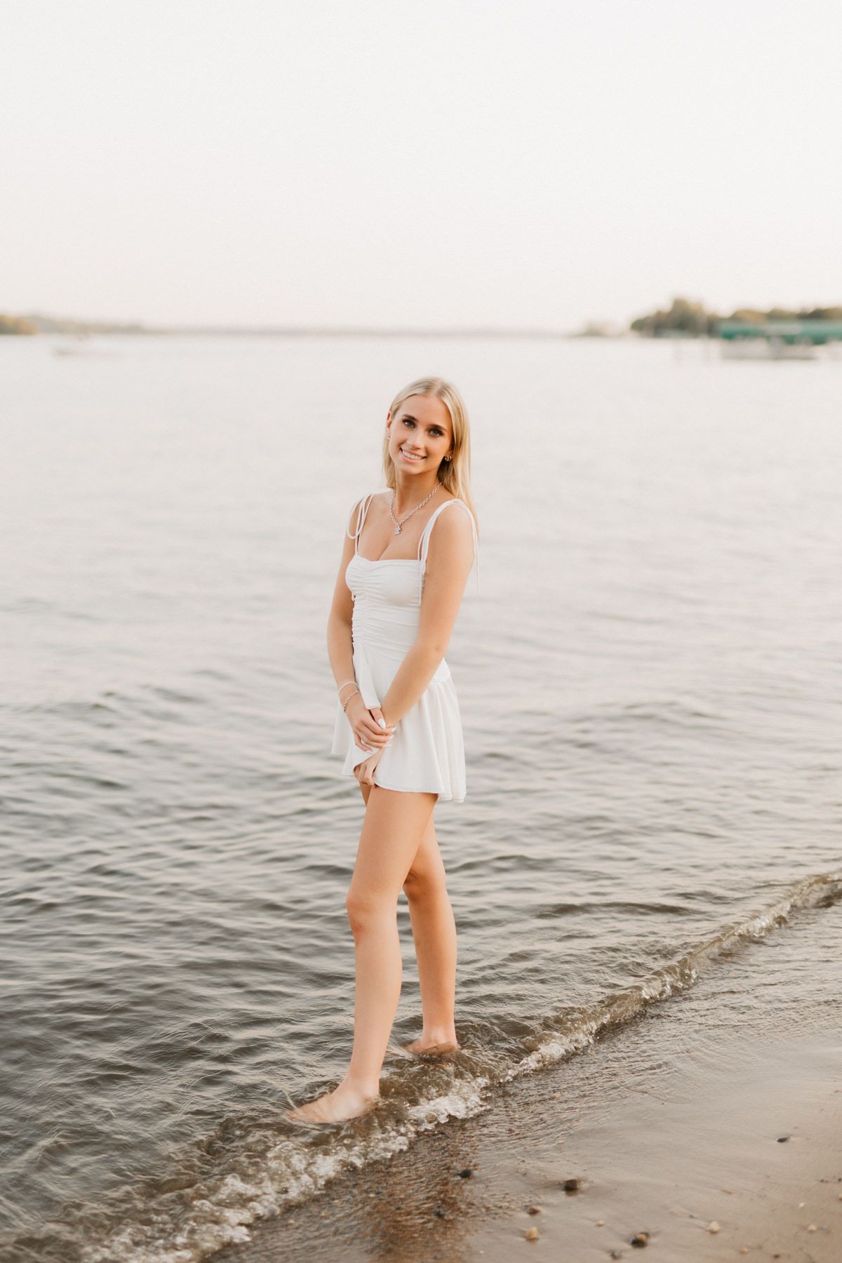 Smiling blonde woman in white sundress standing barefoot at the edge of a calm lake at sunset.