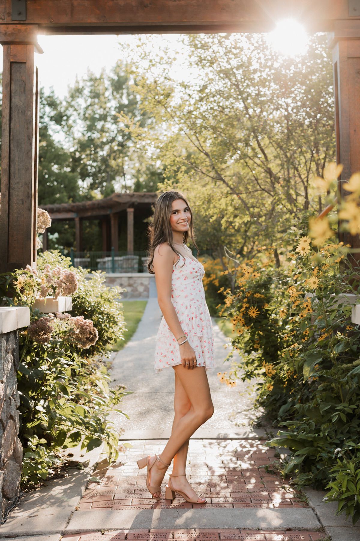 Young woman in floral sundress and heels posing in sunlit garden pathway surrounded by lush greenery.