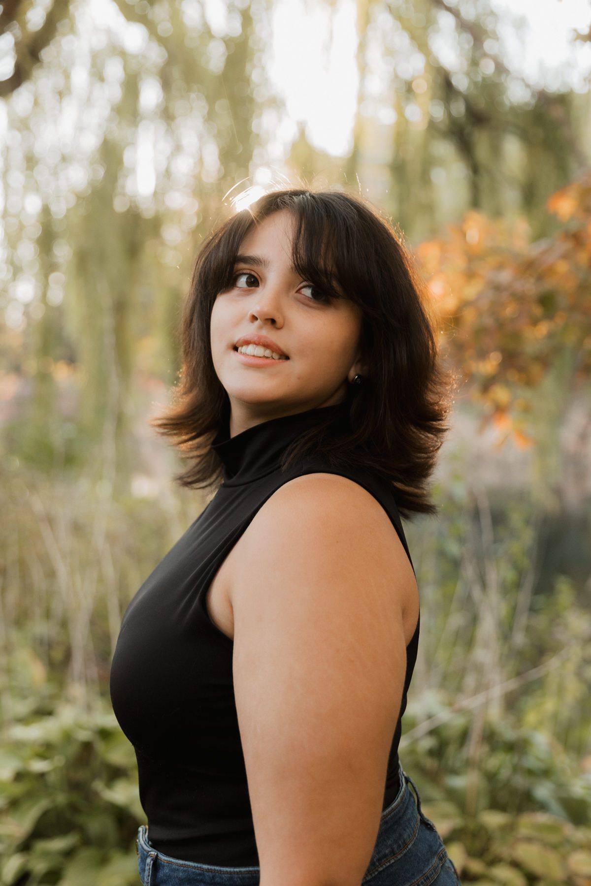 Young brunette woman in black sleeveless turtleneck smiling over shoulder in autumn forest.