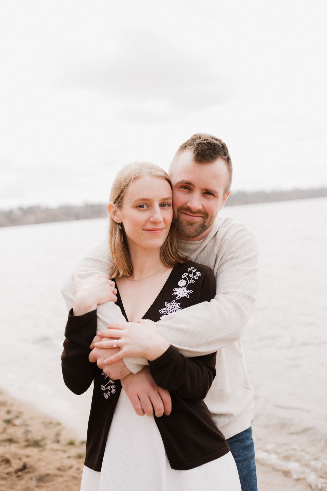 A couple embraces affectionately on a sandy beach shore, the man hugging the woman from behind, both smiling warmly.
