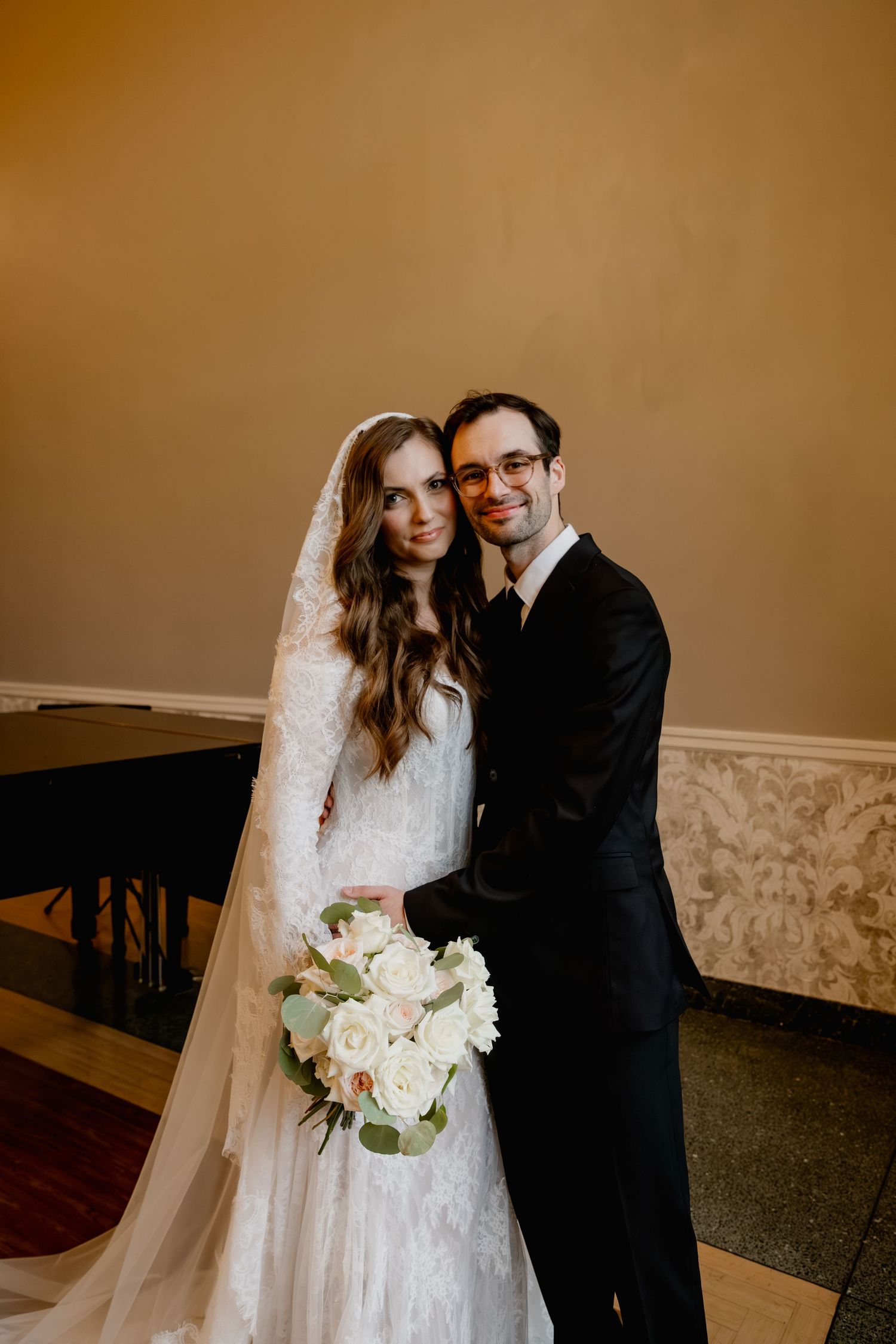 Bride in lace gown with veil holding white rose bouquet stands with groom in black suit for wedding portrait.