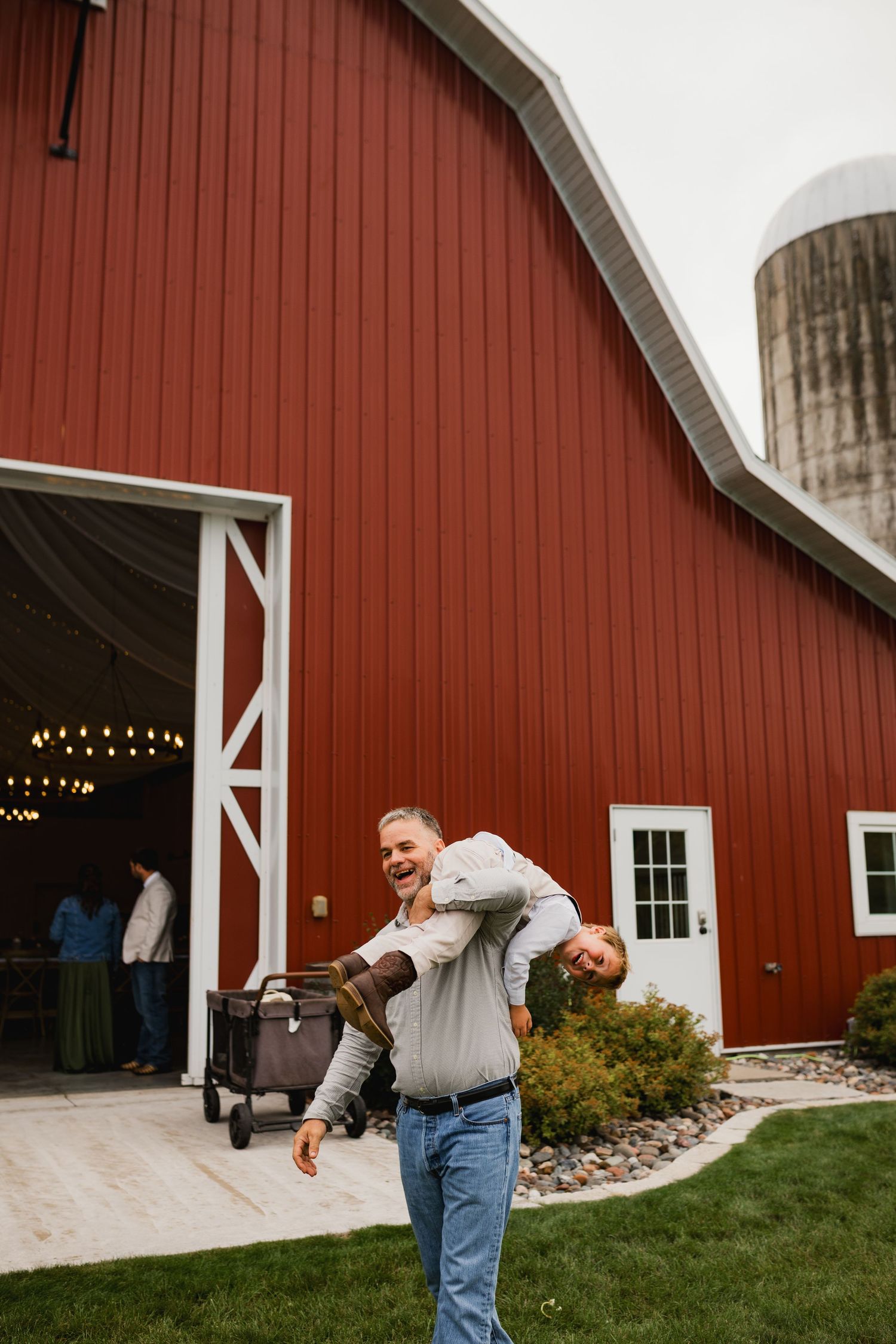 Man giving piggyback ride outside red barn venue with silo, smiling joyfully at rustic countryside event.
