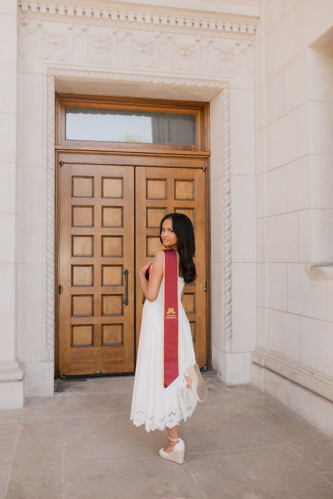Graduate in white dress and maroon stole stands before large wooden double doors of stone building.