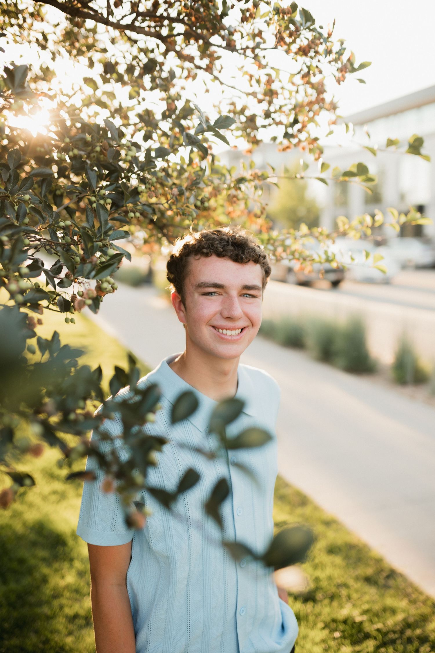 Smiling teenage boy in light blue shirt standing near leafy green tree with golden sunlight in background outdoors.