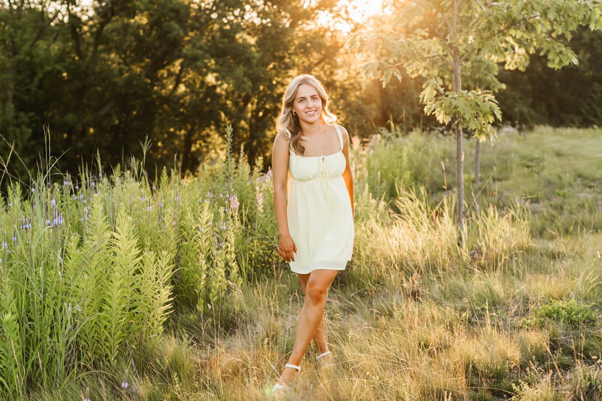 A person in a white summer dress walks through tall grass during golden hour sunset in a natural outdoor setting.