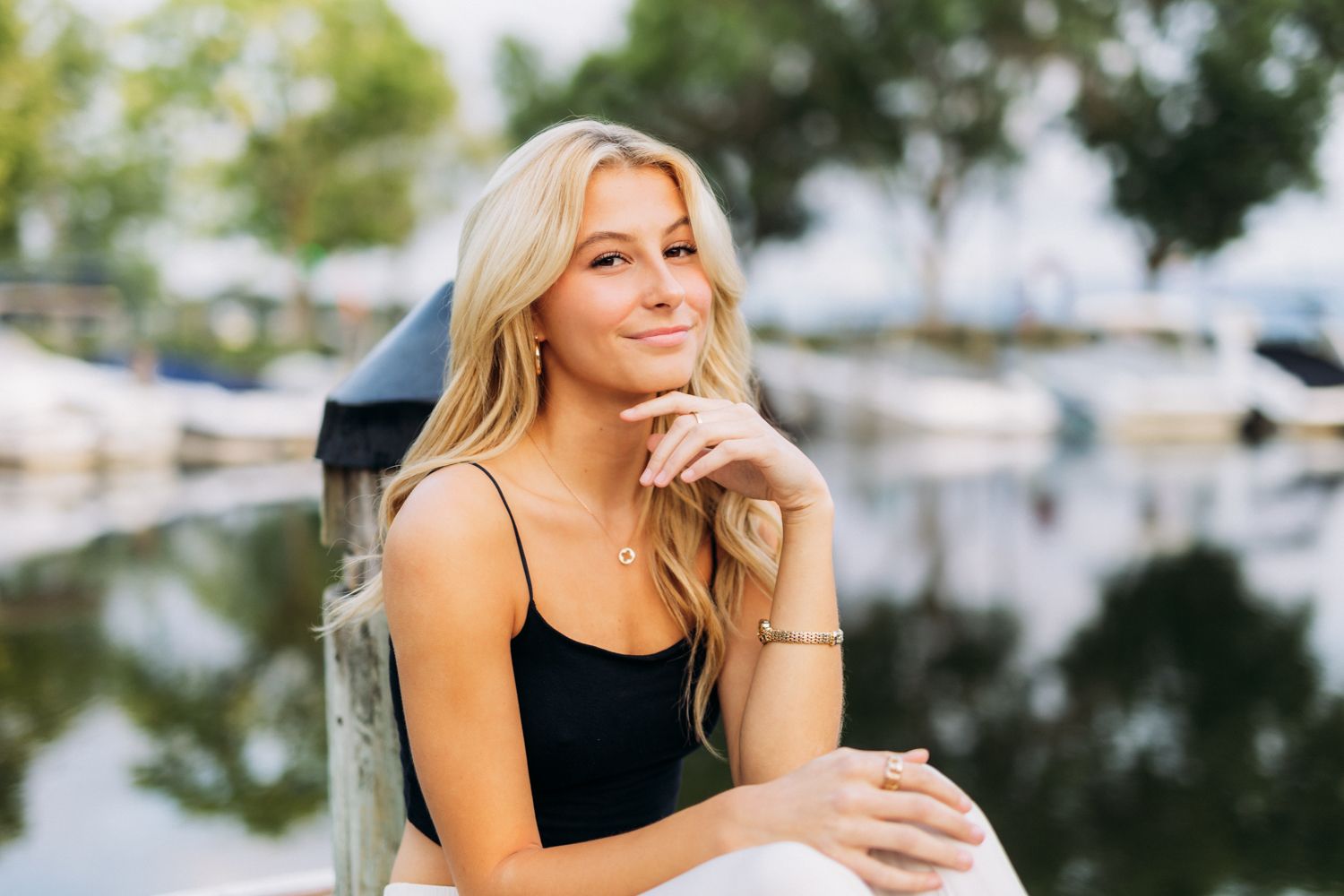 Person with long blonde hair poses thoughtfully by marina with boats and water reflection in soft afternoon light.