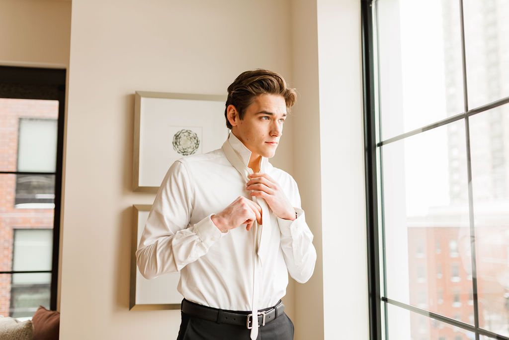Professional in a crisp white shirt poses thoughtfully in front of large windows in minimalist indoor setting.