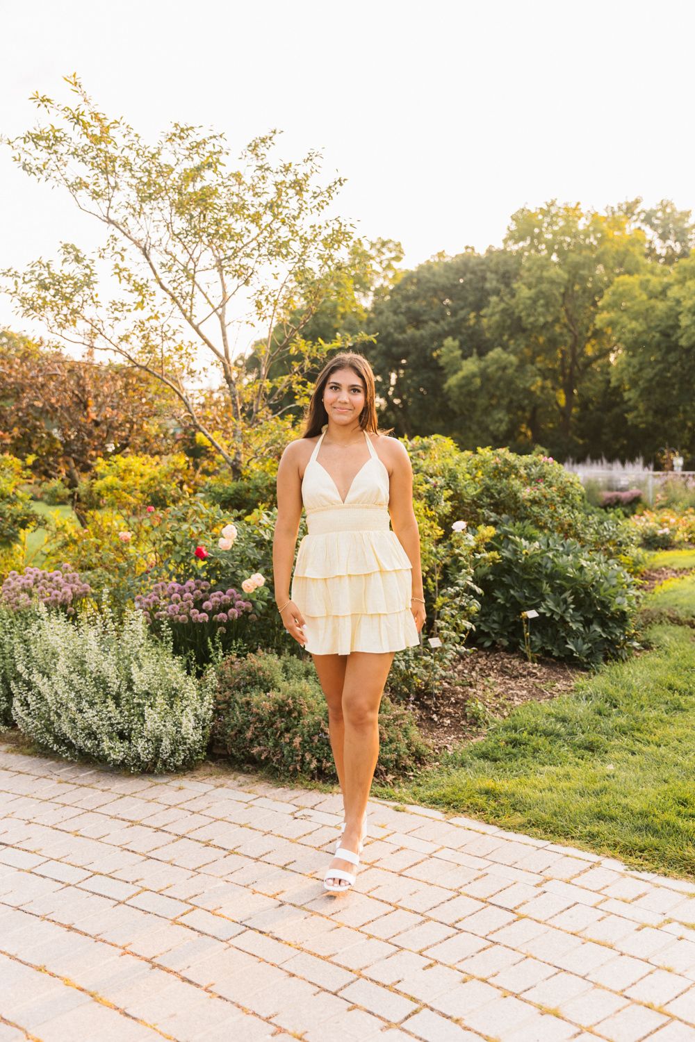 Woman in yellow summer dress poses on stone pathway during golden hour with garden in background.