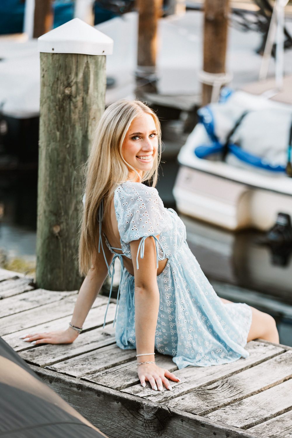 Woman in blue dress poses on wooden dock with boats in background.