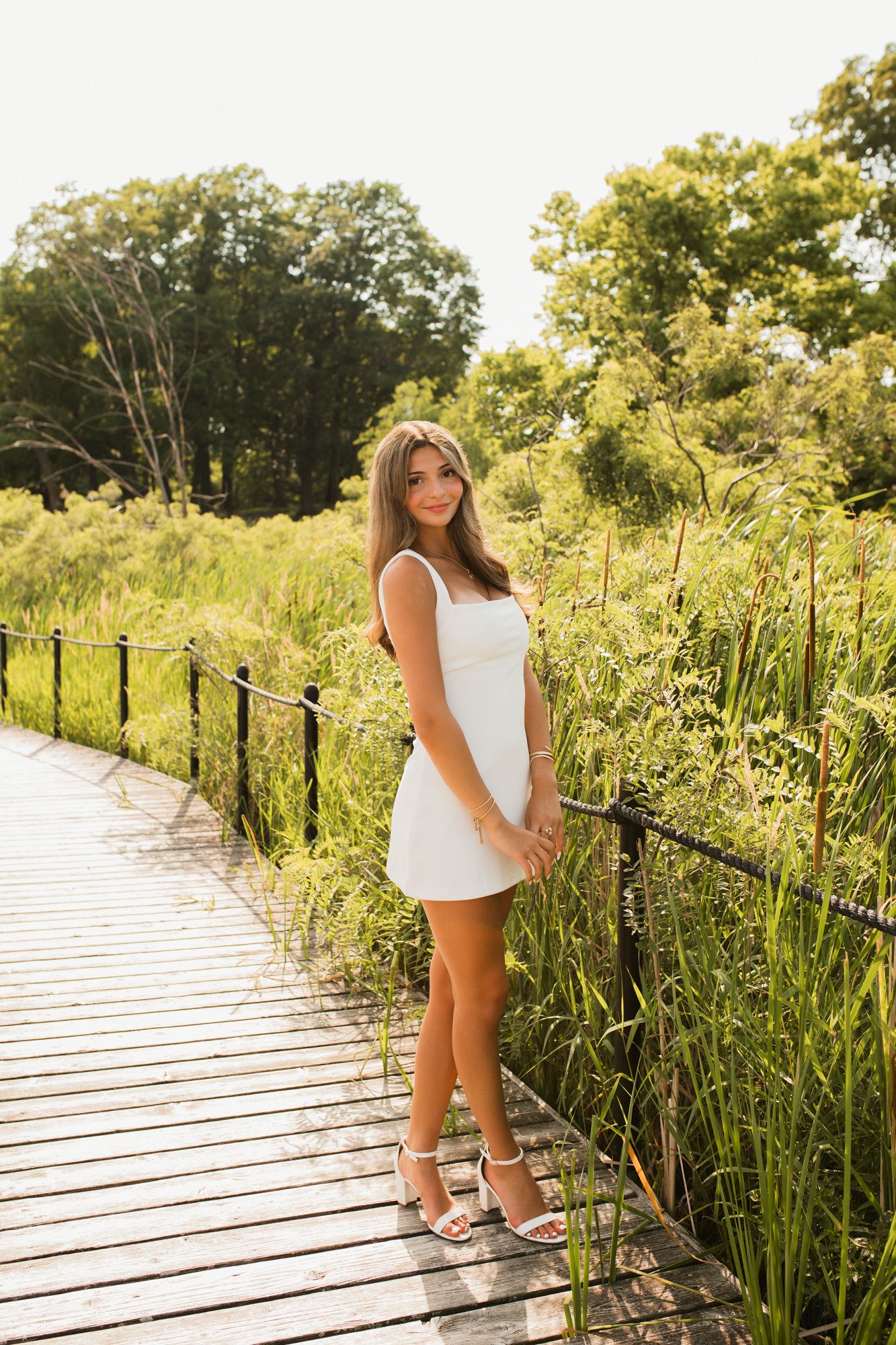 Young woman in white mini dress poses on wooden boardwalk surrounded by lush green reeds and trees on sunny day.