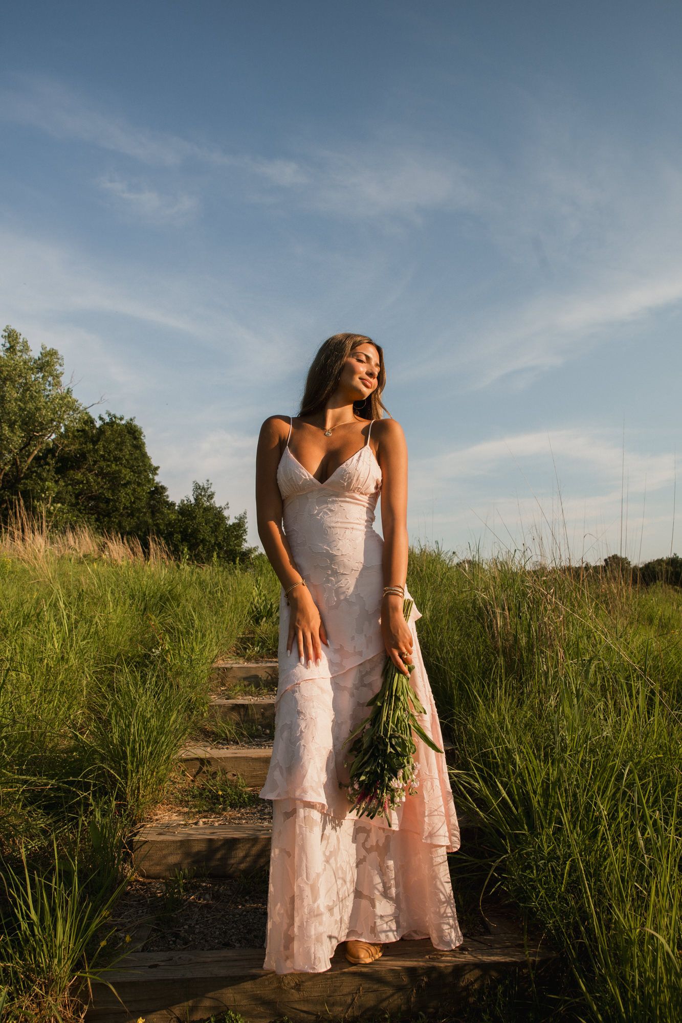Bride in white lace gown holding bouquet on wooden steps in sunlit green field.