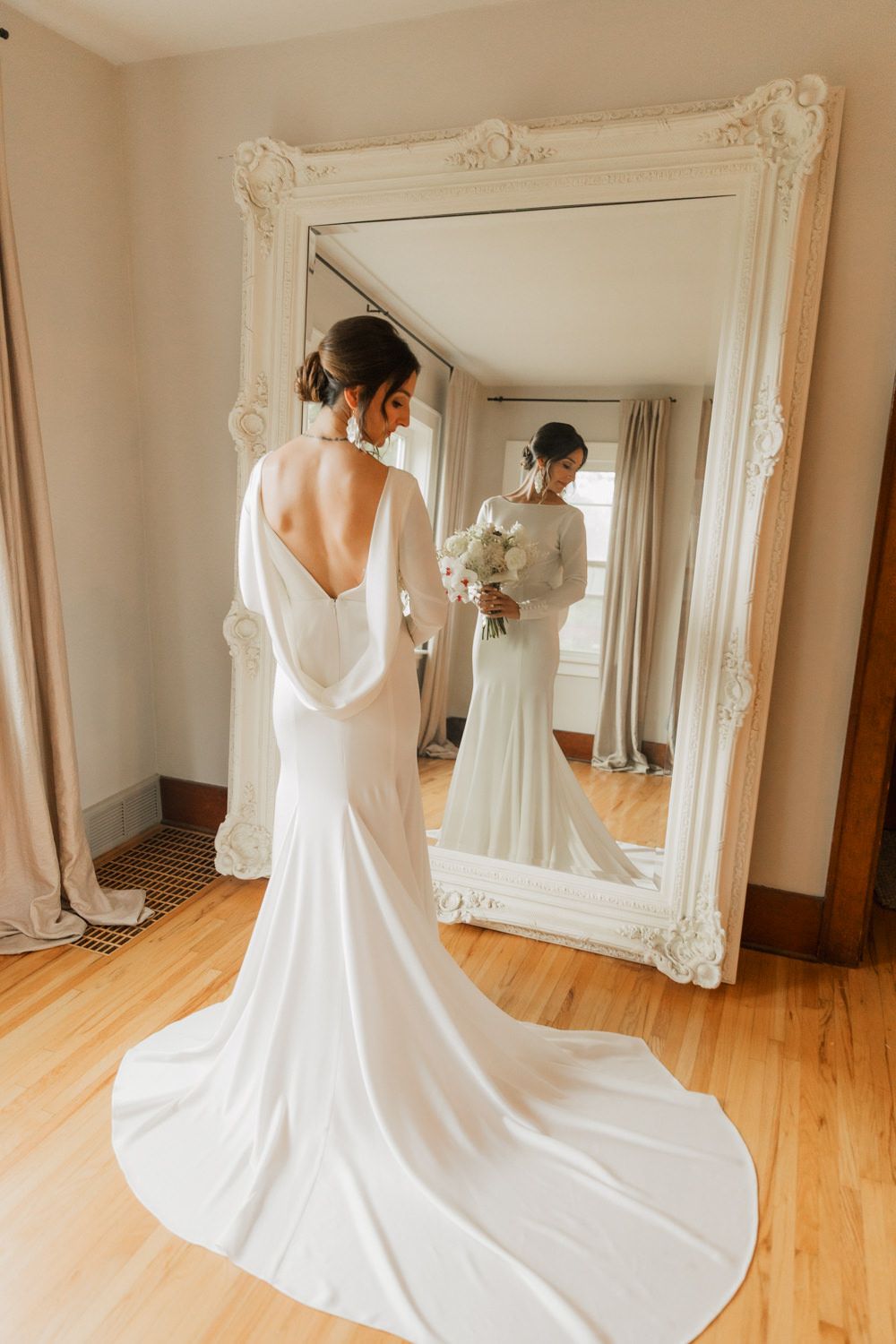 A flowing white wedding dress train spreads elegantly across hardwood floors in front of an ornate mirror.
