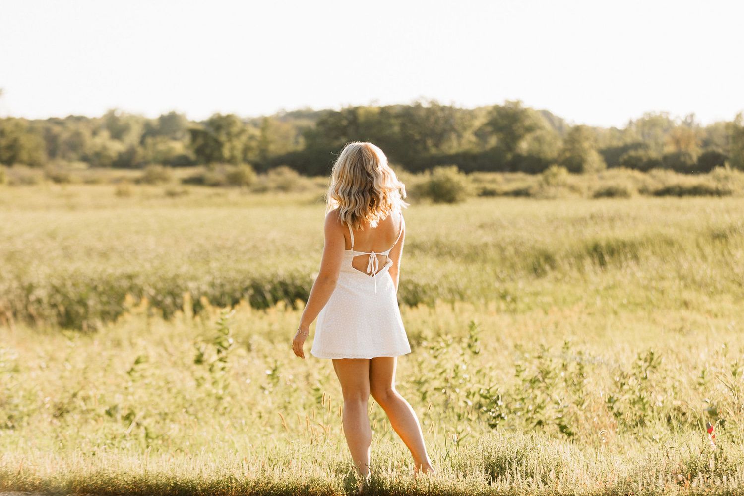 Figure in a white summer dress stands in a sunlit meadow during golden hour.