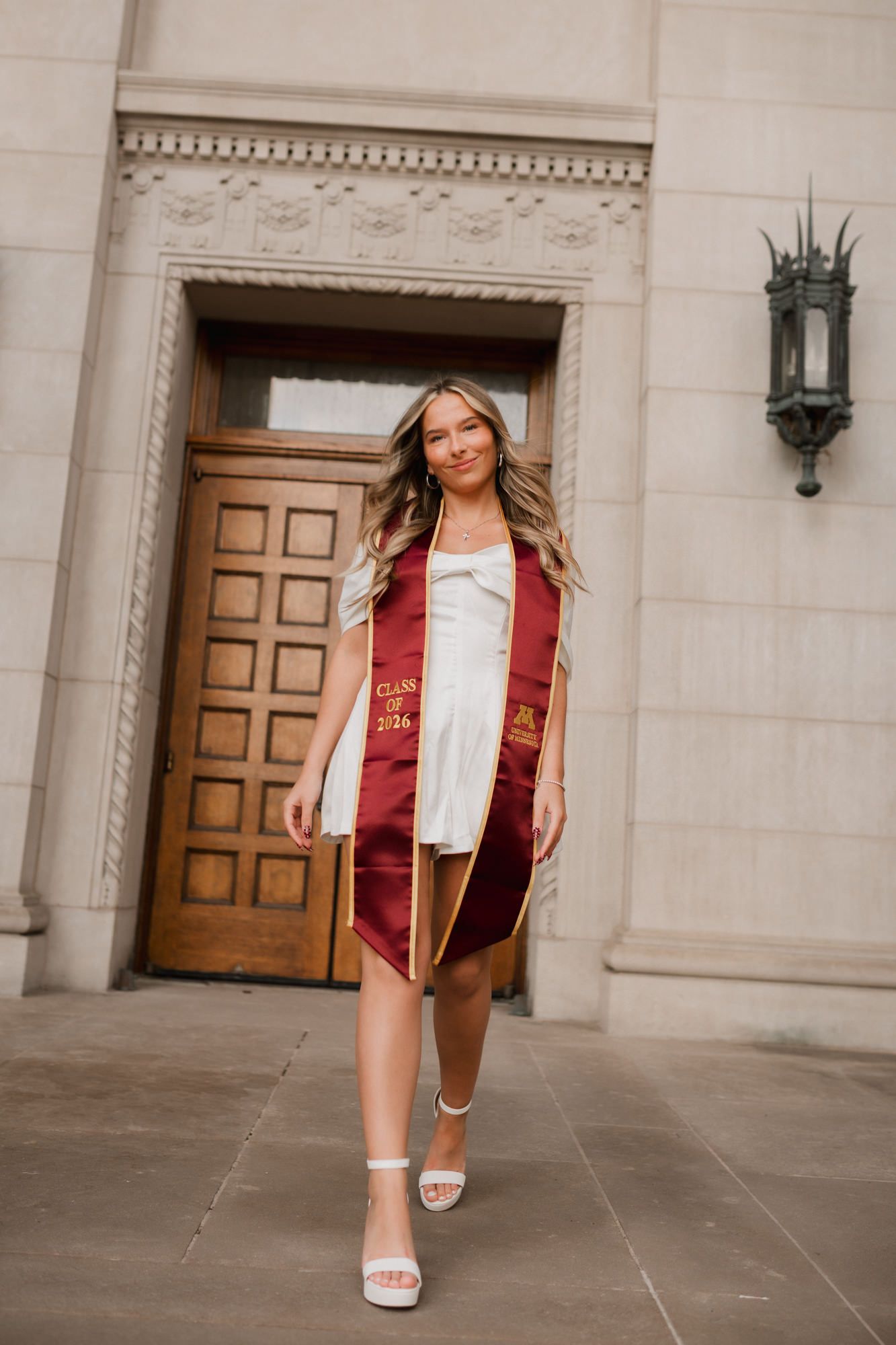 A smiling graduate walks confidently in a white dress and maroon Class of 2024 stole outside an ornate building.