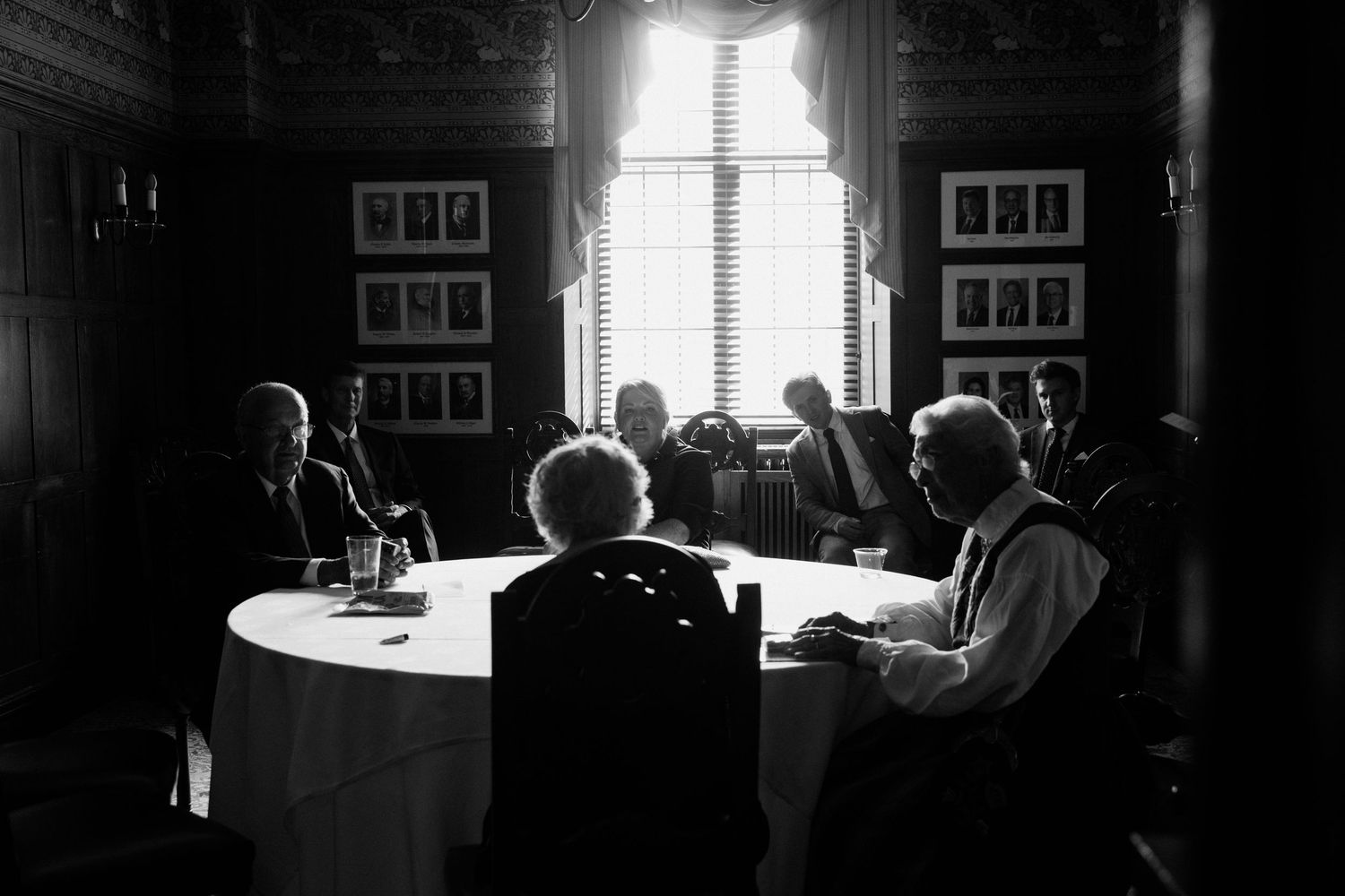 Black and white photo of people seated around a round table in a dark wood-paneled room with framed portraits on the walls.