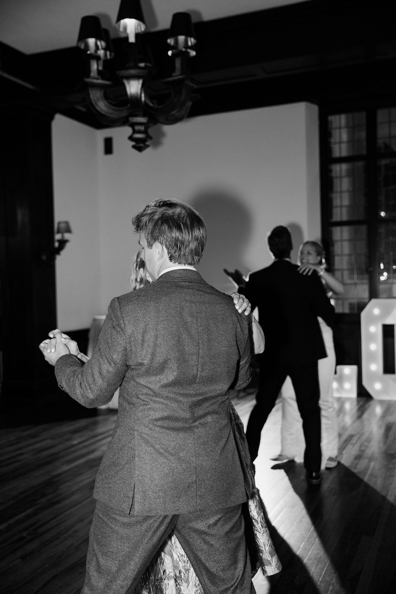 Black and white photo of guests dancing at a wedding reception near large illuminated LOVE letters on the dance floor.
